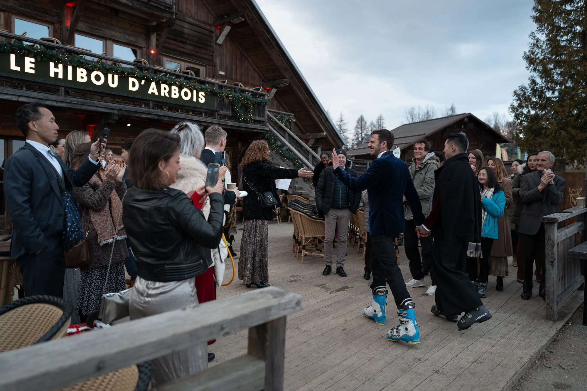 Groupe de personnes souriantes et applaudissant lors d'une célébration en extérieur devant un bâtiment en bois avec le panneau "Le Hibou d'Arbois", certains participant dansant, un homme en fauteuil roulant et un public varié.