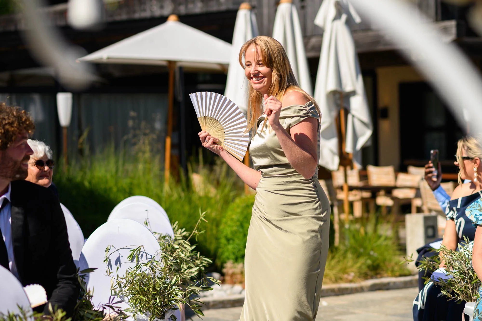 Une femme souriante en robe longue dorée tient un éventail lors d'une cérémonie en plein air, entourée de personnes assises, dont une femme prenant une photo, dans un environnement ensoleillé avec des parasols blancs.