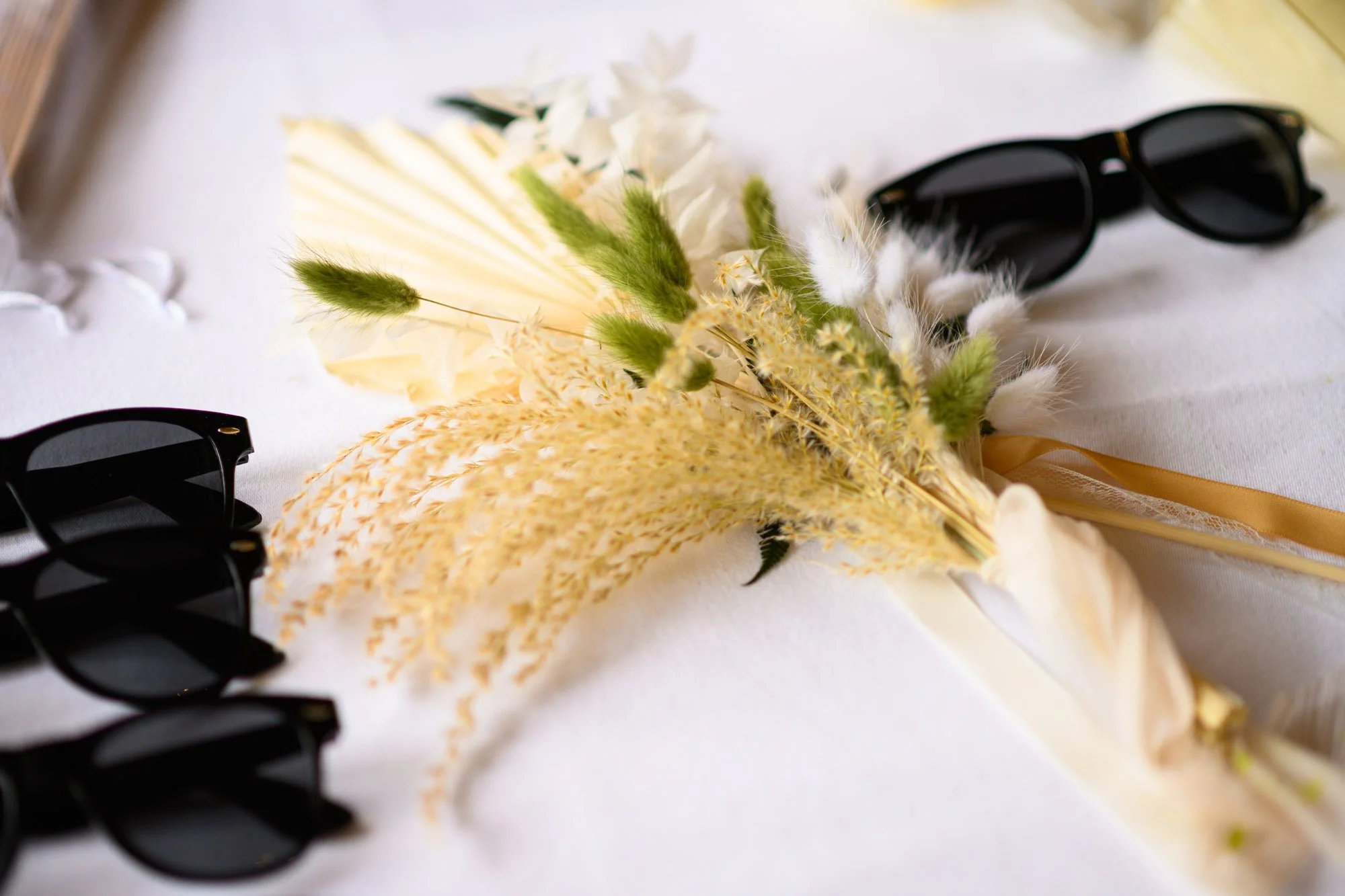 Une paire de lunettes de soleil noires posée sur une surface blanche, accompagnée d'un bouquet de fleurs séchées de couleurs pastel avec du vert, dans un style naturaliste et épuré.