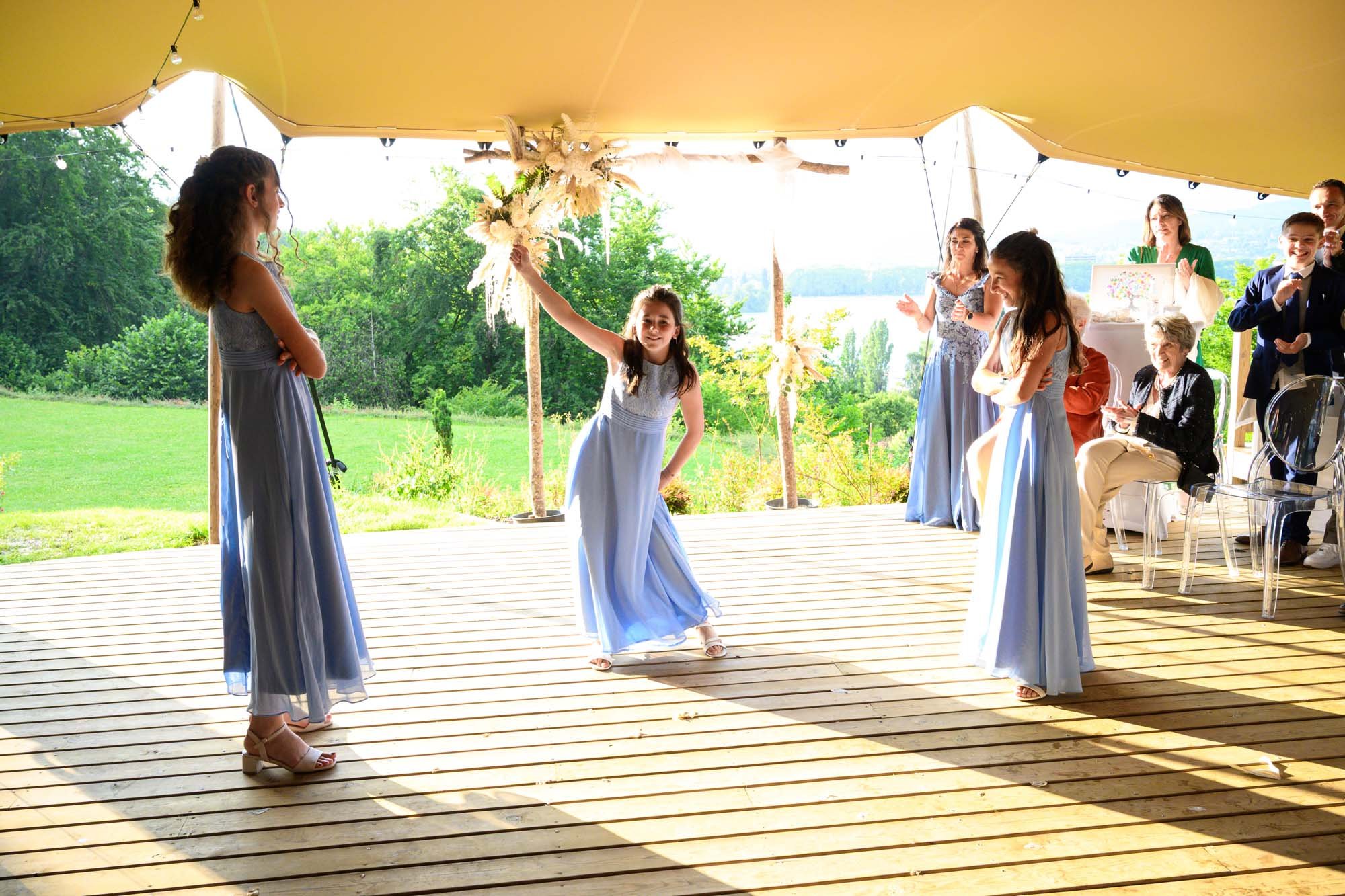 Une jeune fille danse lors d'une fête en plein air, sous un grand chapiteau jaune, avec d'autres enfants et adultes qui applaudissent et regardent, entourés de verdure et d'un paysage en arrière-plan.