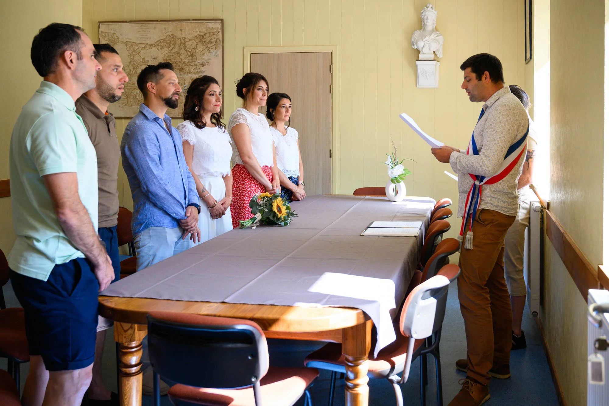 Un groupe de personnes se tenant debout devant un homme qui lit un document, dans une salle avec une table recouverte d'une nappe, décorée de fleurs, avec un tableau sur le mur en arrière-plan.