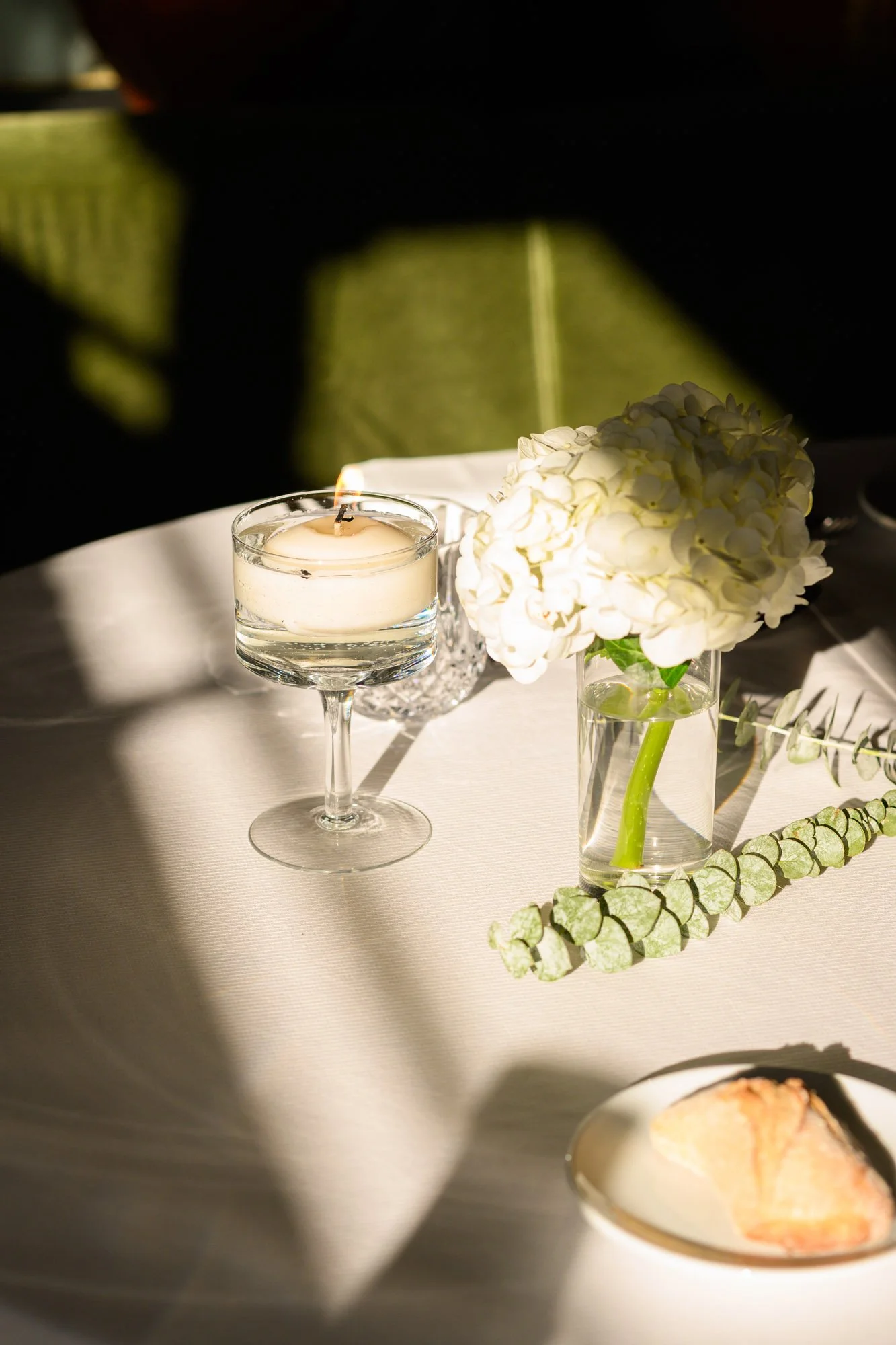 Table avec une bougie allumée, un vase avec un bouquet de hortensias blancs, une plante en feuille verte et une assiette avec un morceau de gâteau, éclairé par la lumière du soleil.