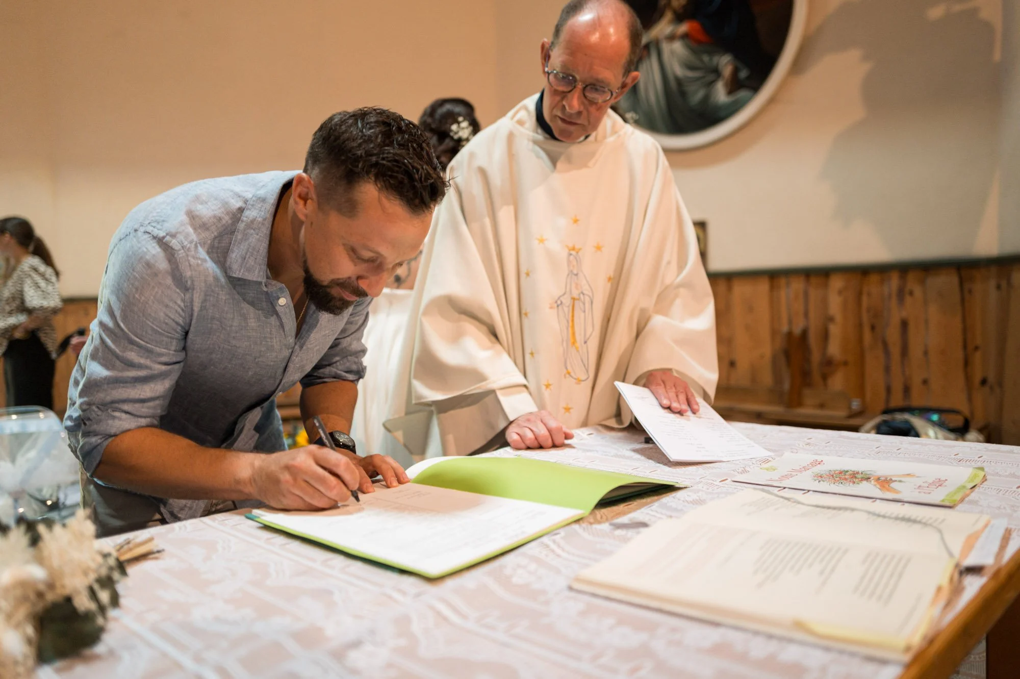 Un homme signe un livre sur une table décorée, un prêtre en robe blanche se tient à côté, dans une église, lors d'une cérémonie.
