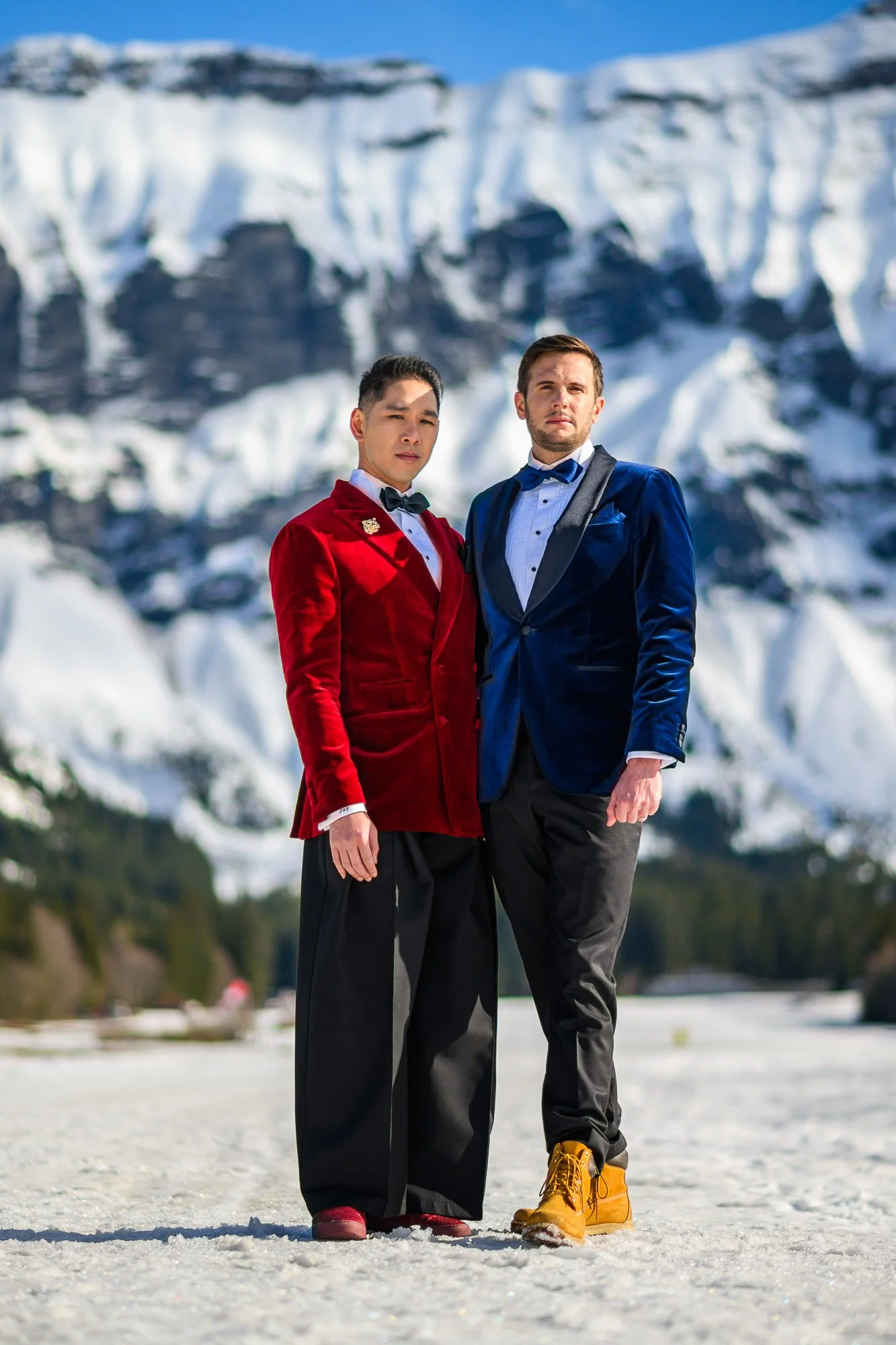 Deux hommes en costumes élégants debout dans un paysage enneigé avec des montagnes en arrière-plan.