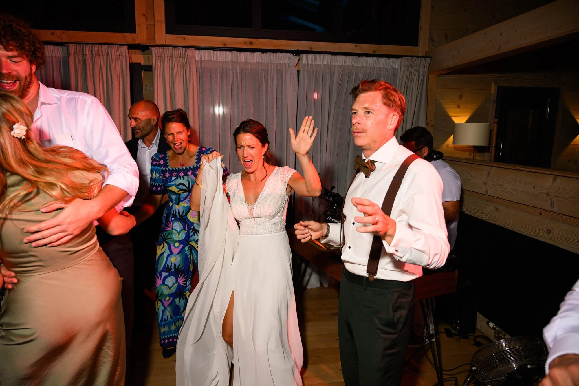 Groupe de personnes dansant et souriant lors d'une fête ou d'un mariage dans une pièce en bois, ambiance festive.
