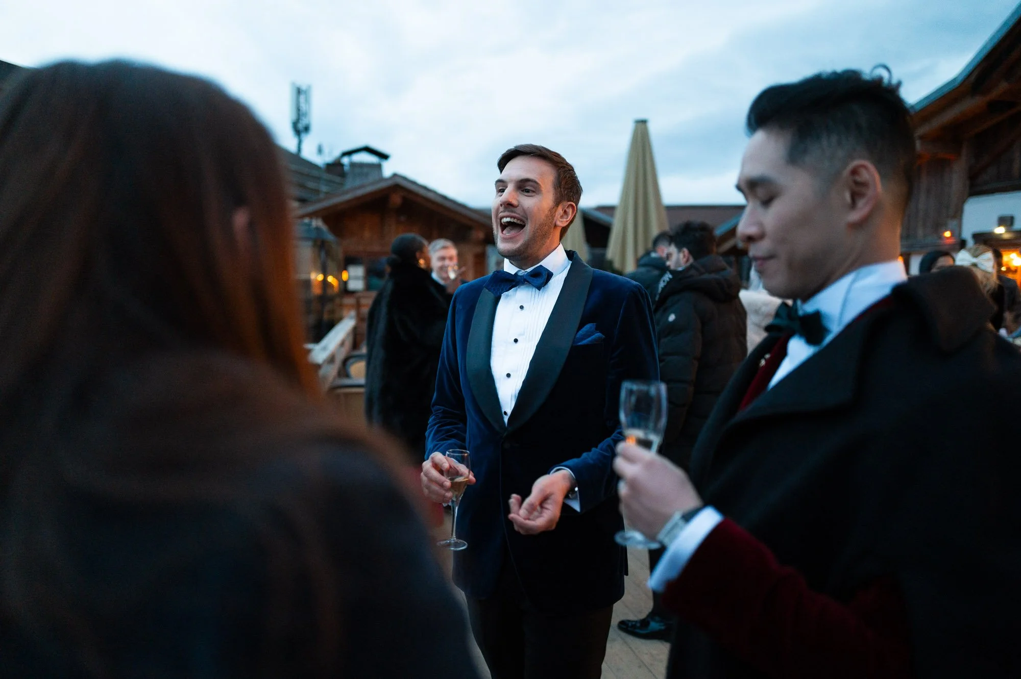 Groupe de personnes lors d'une réception ou fête en plein air, habillées en tenue formelle, discutant et tenant des verres de champagne, le ciel est en début de soirée.