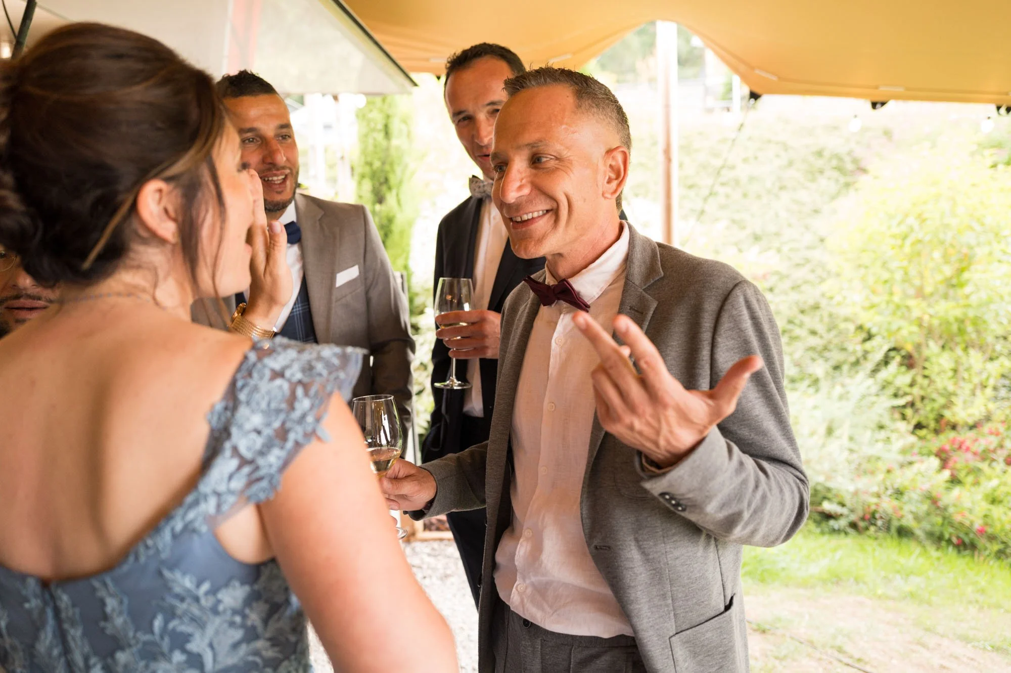 Groupe de personnes en costume discutant à un événement en plein air, avec un homme en train de parler et de sourire, tout en tenant un verre de vin.