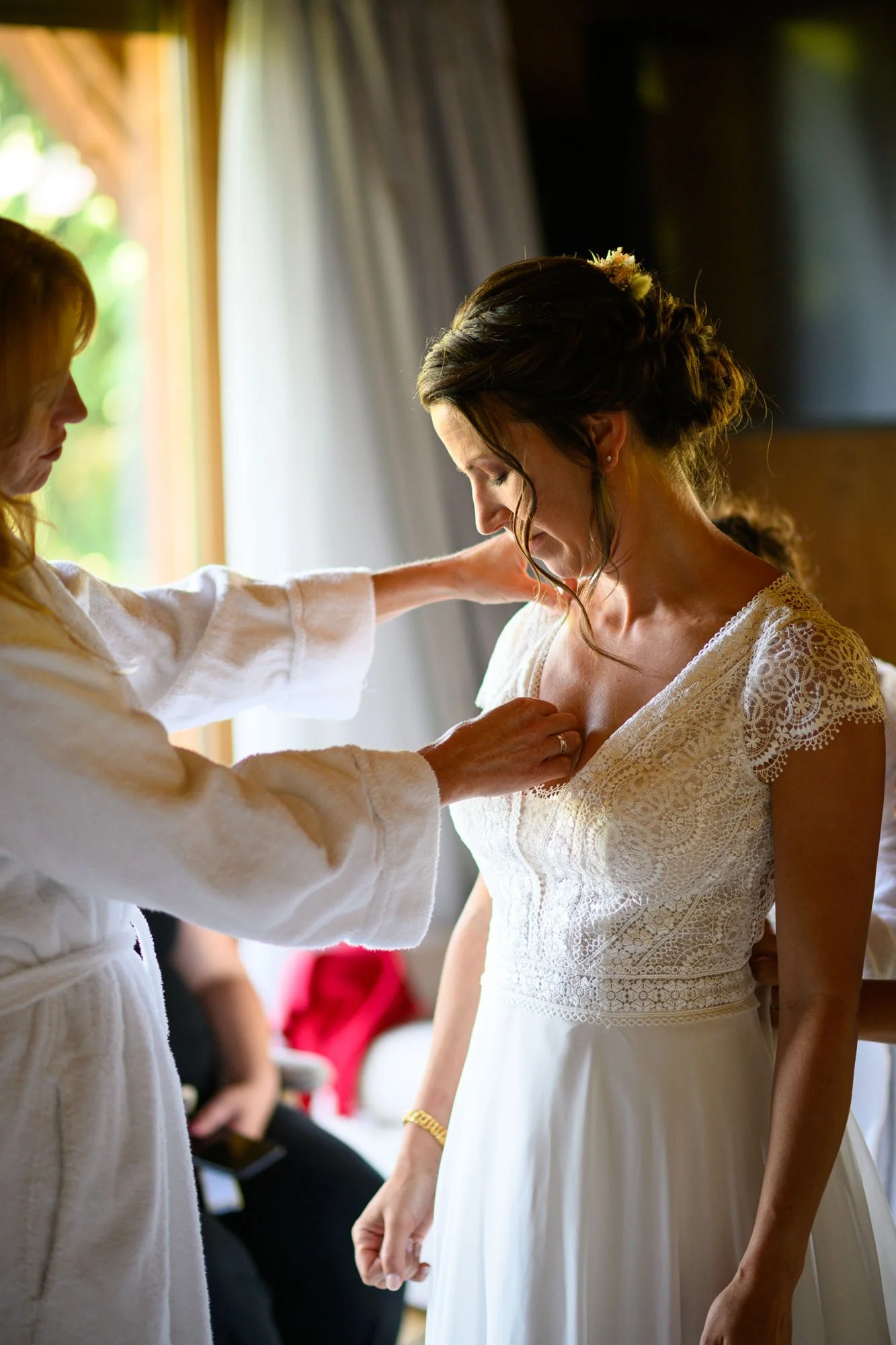 Une femme en robe de mariée reçoit de l'aide pour ajuster son vêtement après une touche finale, dans une pièce lumineuse avec une fenêtre en arrière-plan.