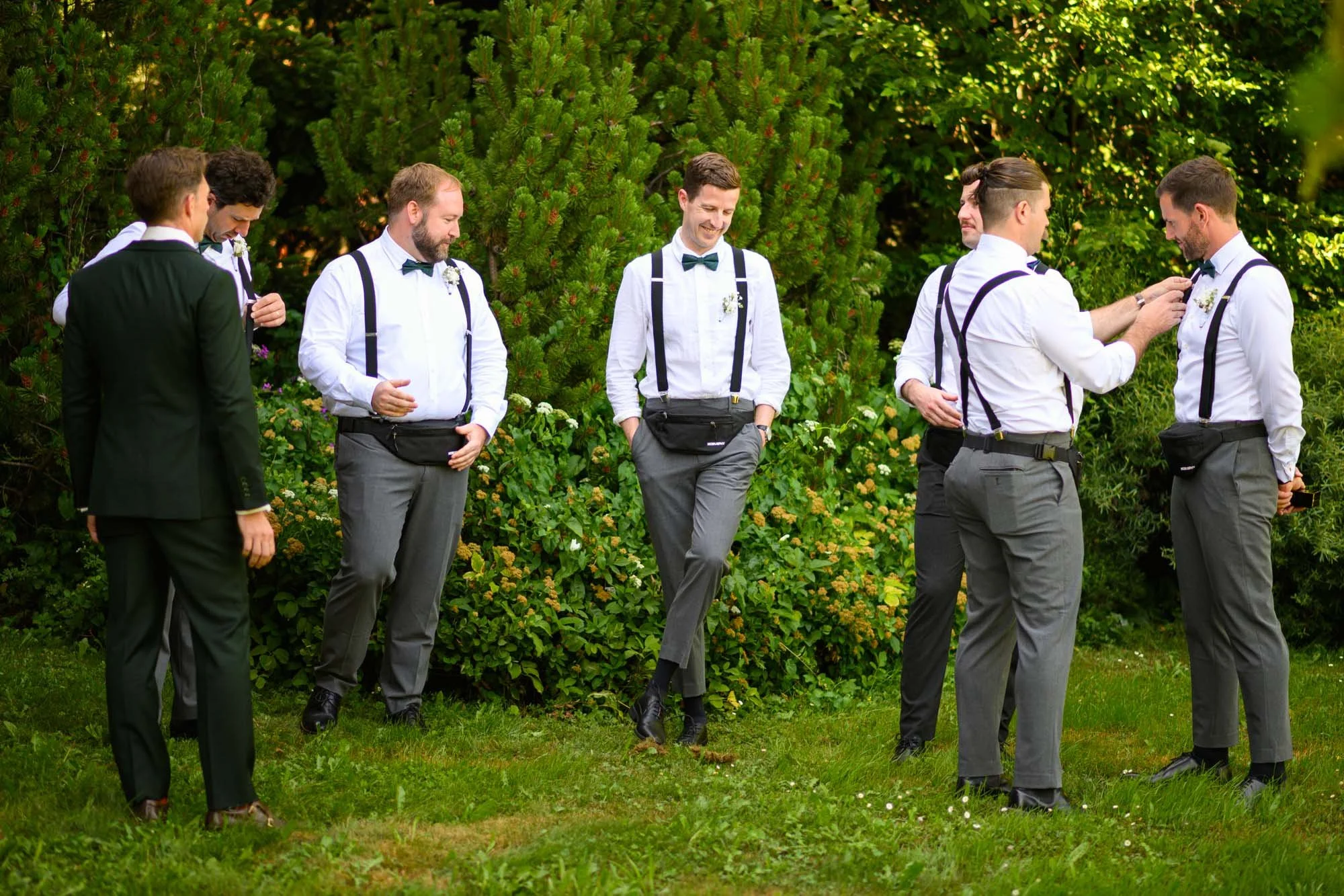 Groupe de jeunes hommes en tenue de mariage, certains ajustant des accessoires, dans un jardin verdoyant