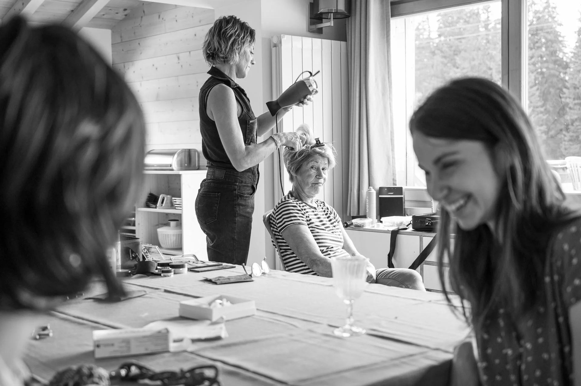 Une femme se fait coiffer par une autre femme dans une pièce lumineuse avec vue sur l'extérieur, d'autres femmes souriantes sont assises autour d'une table.