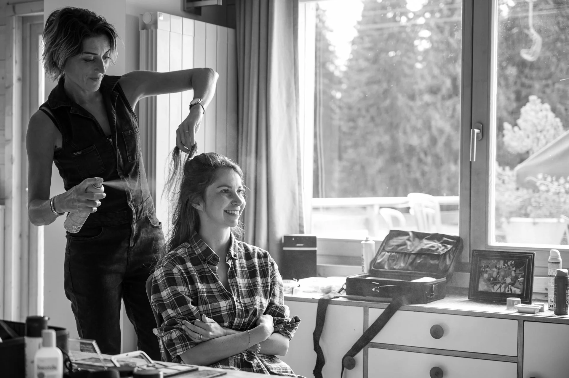 Une femme se fait coiffer par une autre dans une pièce lumineuse, avec une grande fenêtre derrière.
