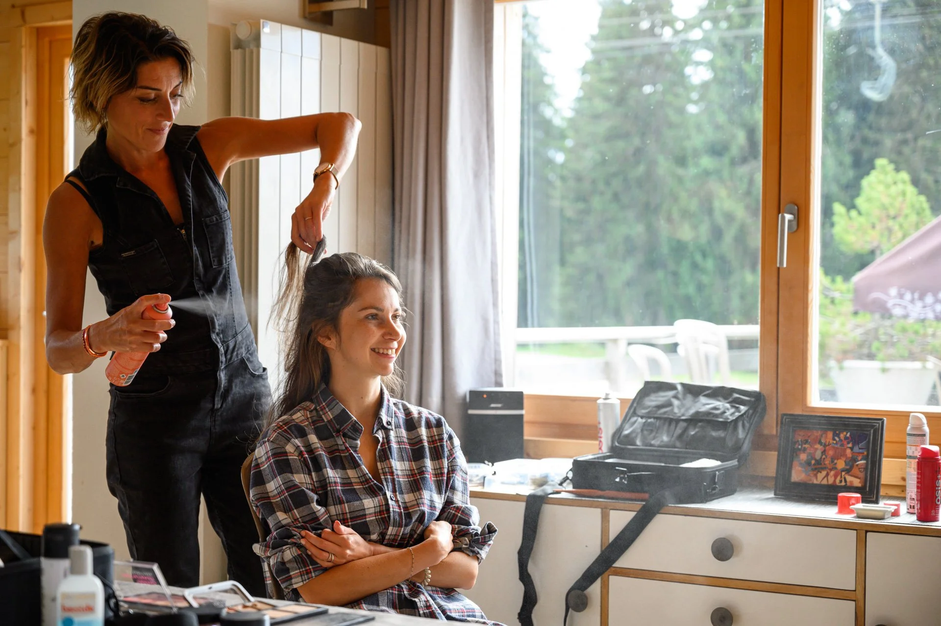 Une femme coiffe une autre femme dans une pièce lumineuse avec vue sur la nature.