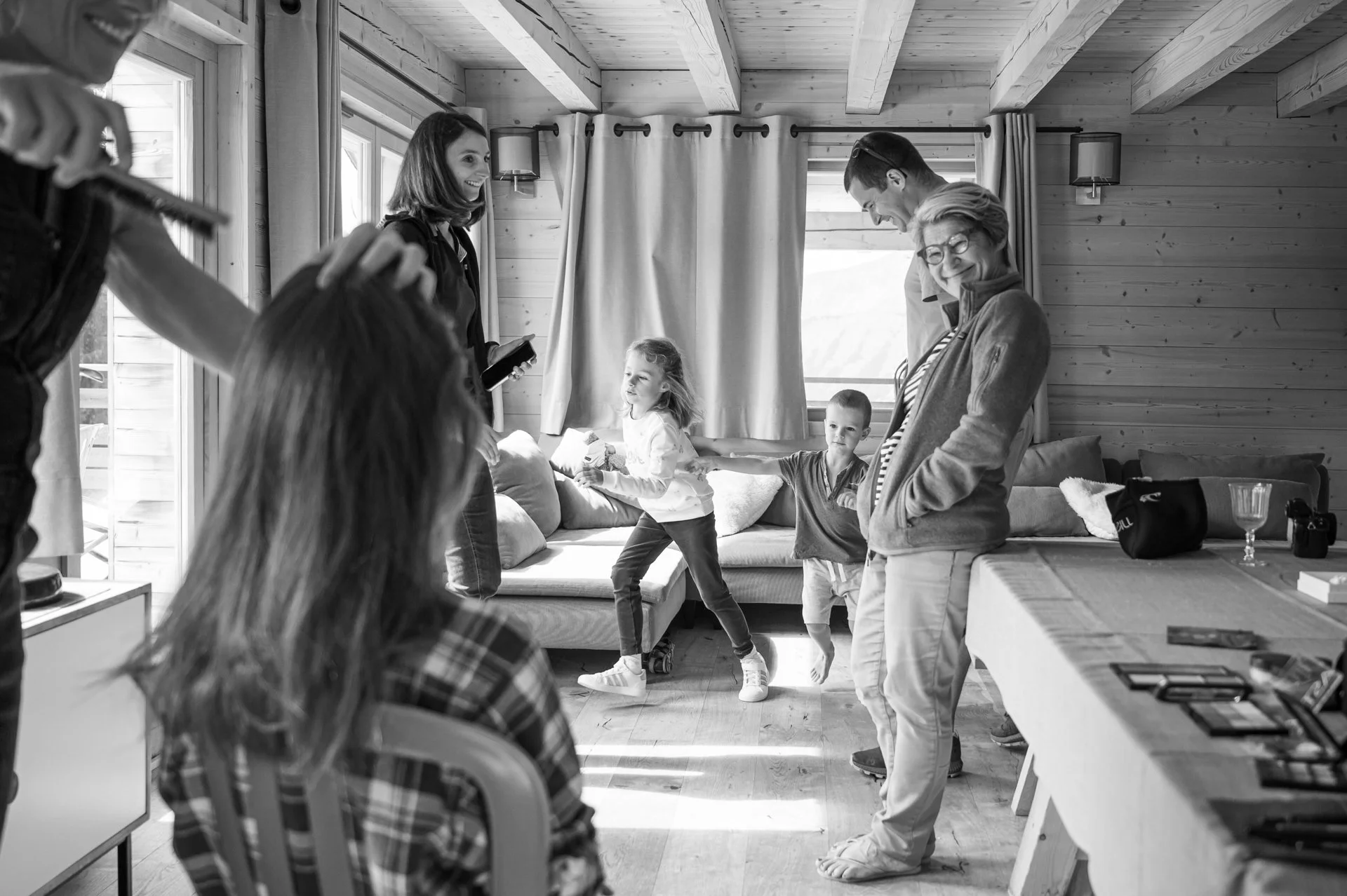 Une famille et des amis qui s'amusent dans un salon en bois, certains dansent et d'autres regardent, avec des grandes fenêtres et un décor cosy.