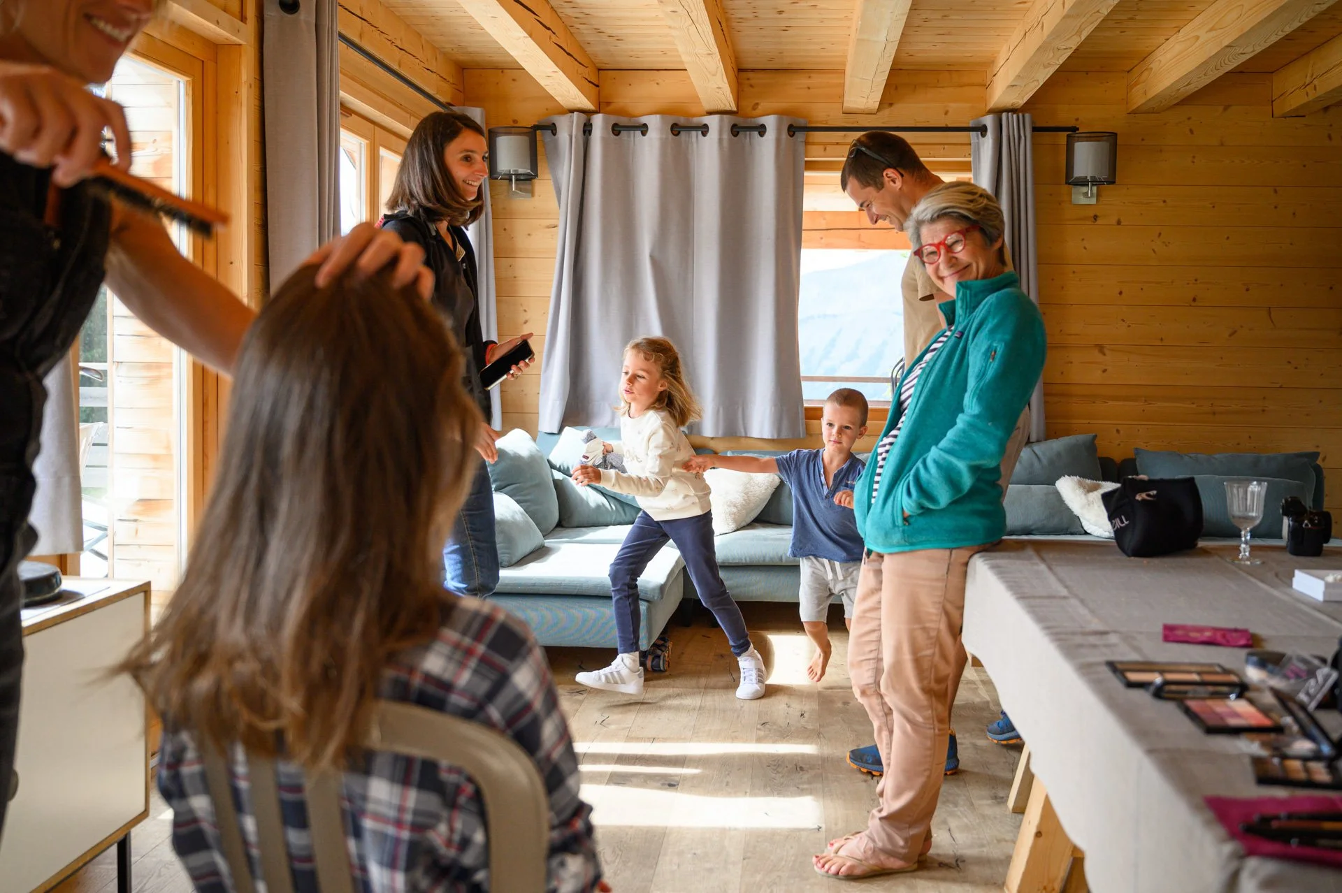 Une famille et quelques amis sont dans un salon en bois, certains sont assis, d'autres jouent ou discutent. Il y a des jeunes et une femme âgée avec des lunettes rouges, souriant.