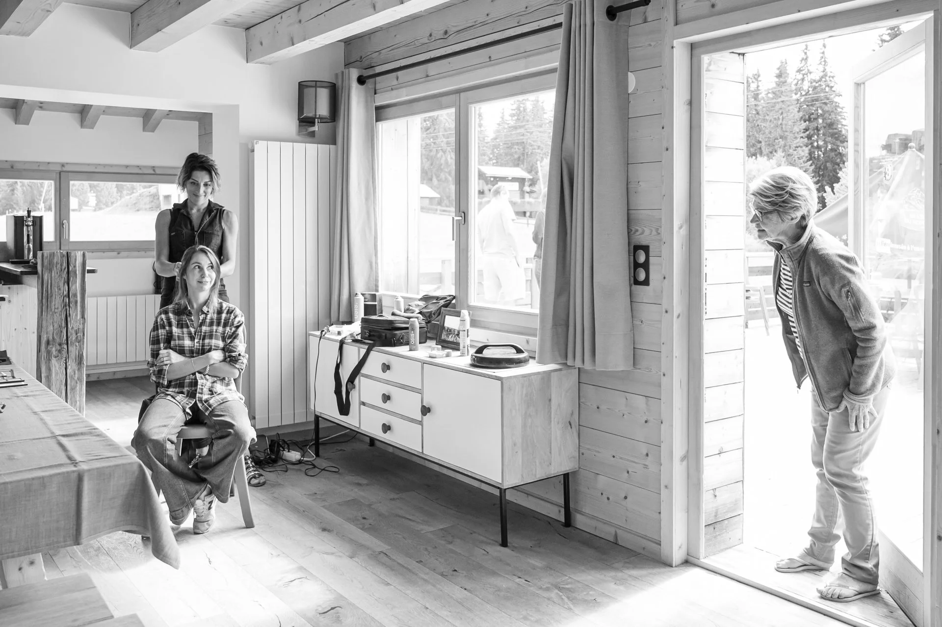 Une femme plus âgée qui salonne deux jeunes femmes à l'entrée d'une maison en bois, à l'intérieur. Il y a une femme assise sur une chaise, une autre debout derrière elle, et une autre femme appelée à l'extérieur.