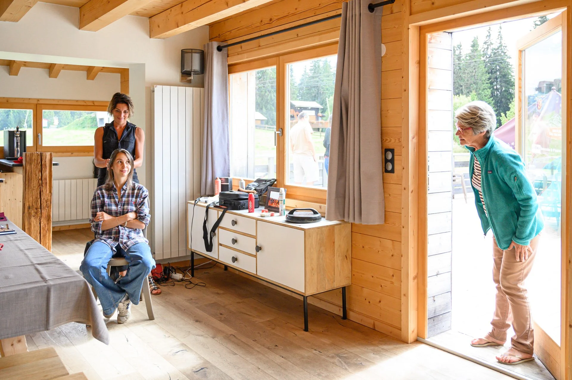 trois femmes dans une maison en bois avec de grandes fenêtres, une femme est assise et l'autre derrière elle, toutes deux près d'une table, une autre femme à la porte en flip-flops, une vue extérieure avec des arbres