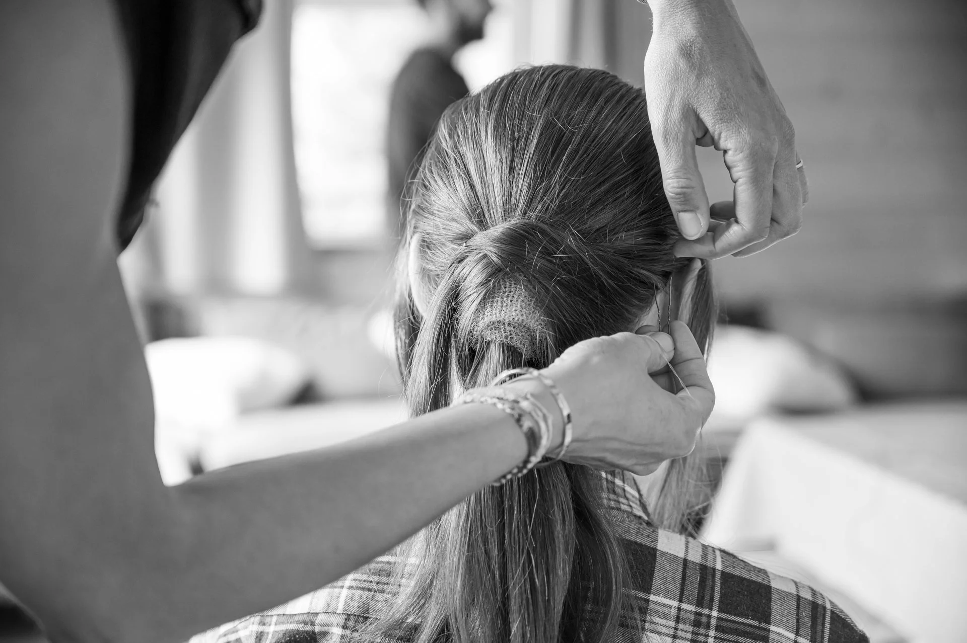 Une personne coiffe une autre personne, probablement une perruque ou des cheveux attachés, dans une pièce avec lumière naturelle.
