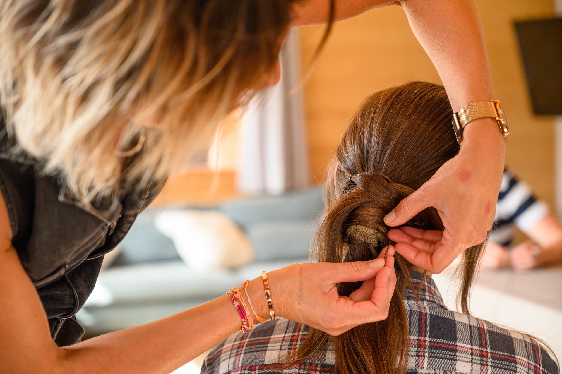 Une femme coiffe une autre personne, en tressant ses cheveux, dans un intérieur chaleureux.