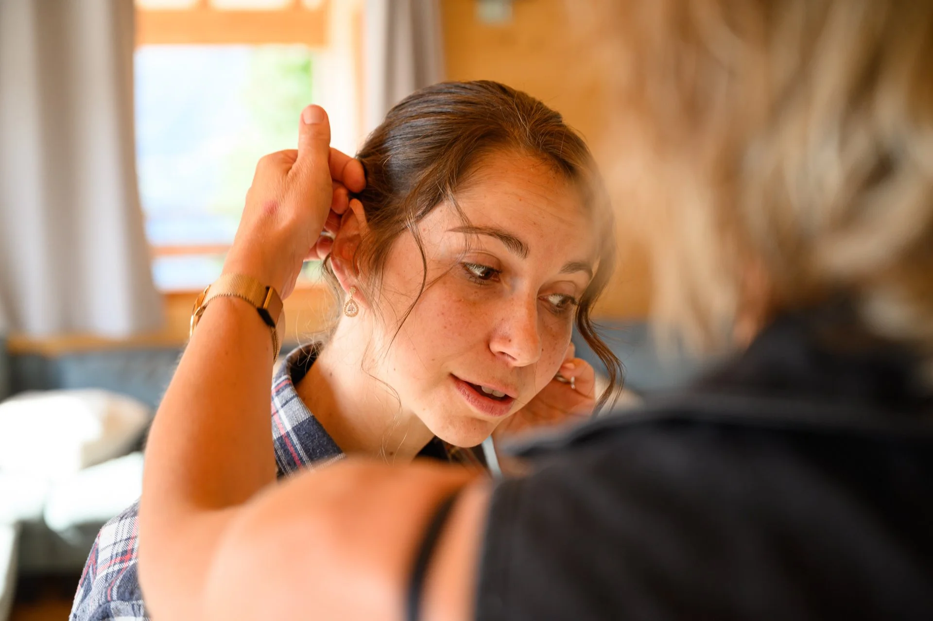 Une jeune femme aux cheveux bruns, portant une montre dorée et des boucles d'oreilles, ajuste ses cheveux alors qu'elle parle avec une autre personne, dans un intérieur chaleureux et lumineux.
