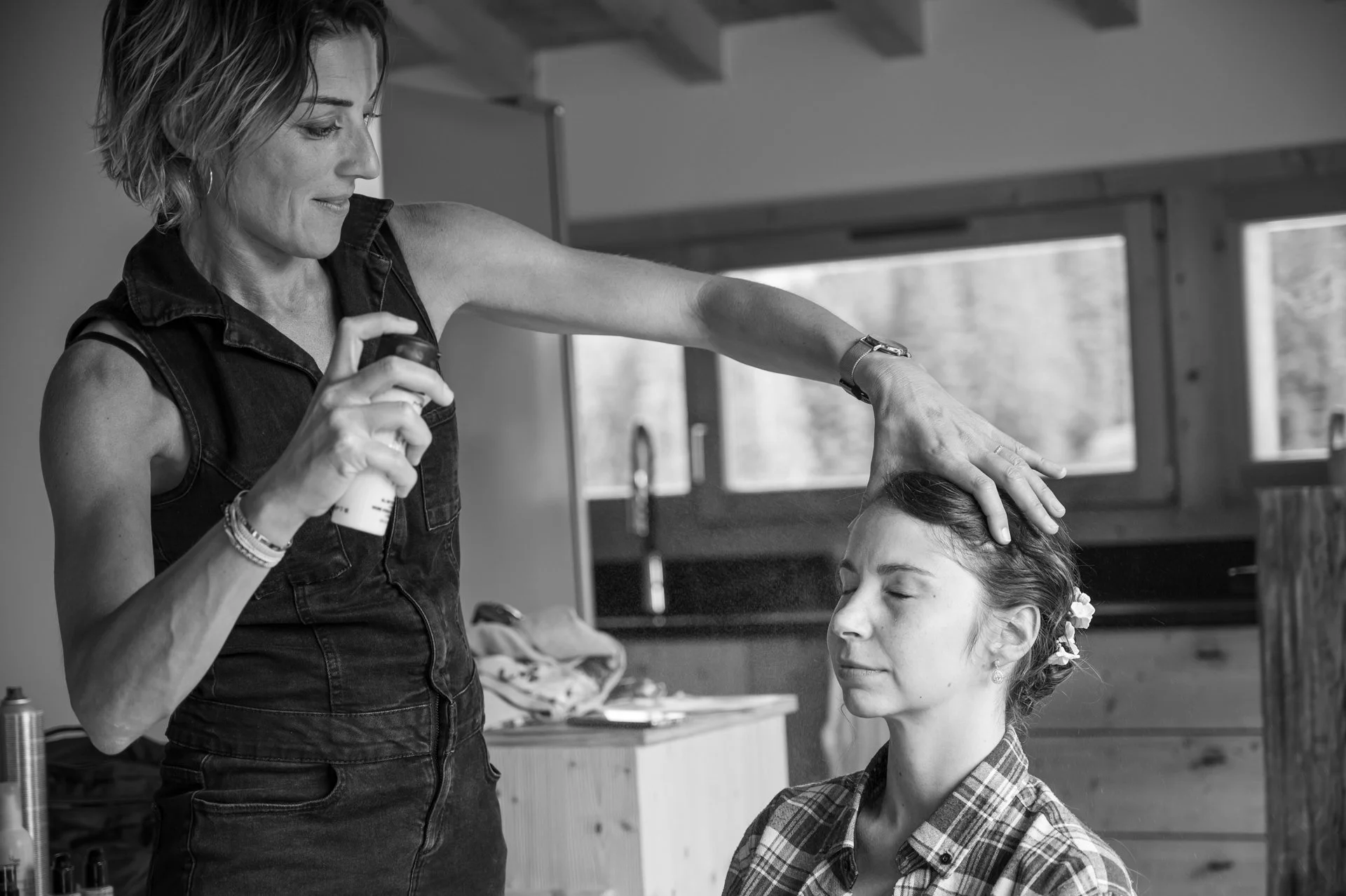 Une femme maquille ou coiffe une jeune fille dans un environnement intérieur avec des fenêtres. La femme tient un spray et touche la tête de la jeune fille, qui a les yeux fermés.