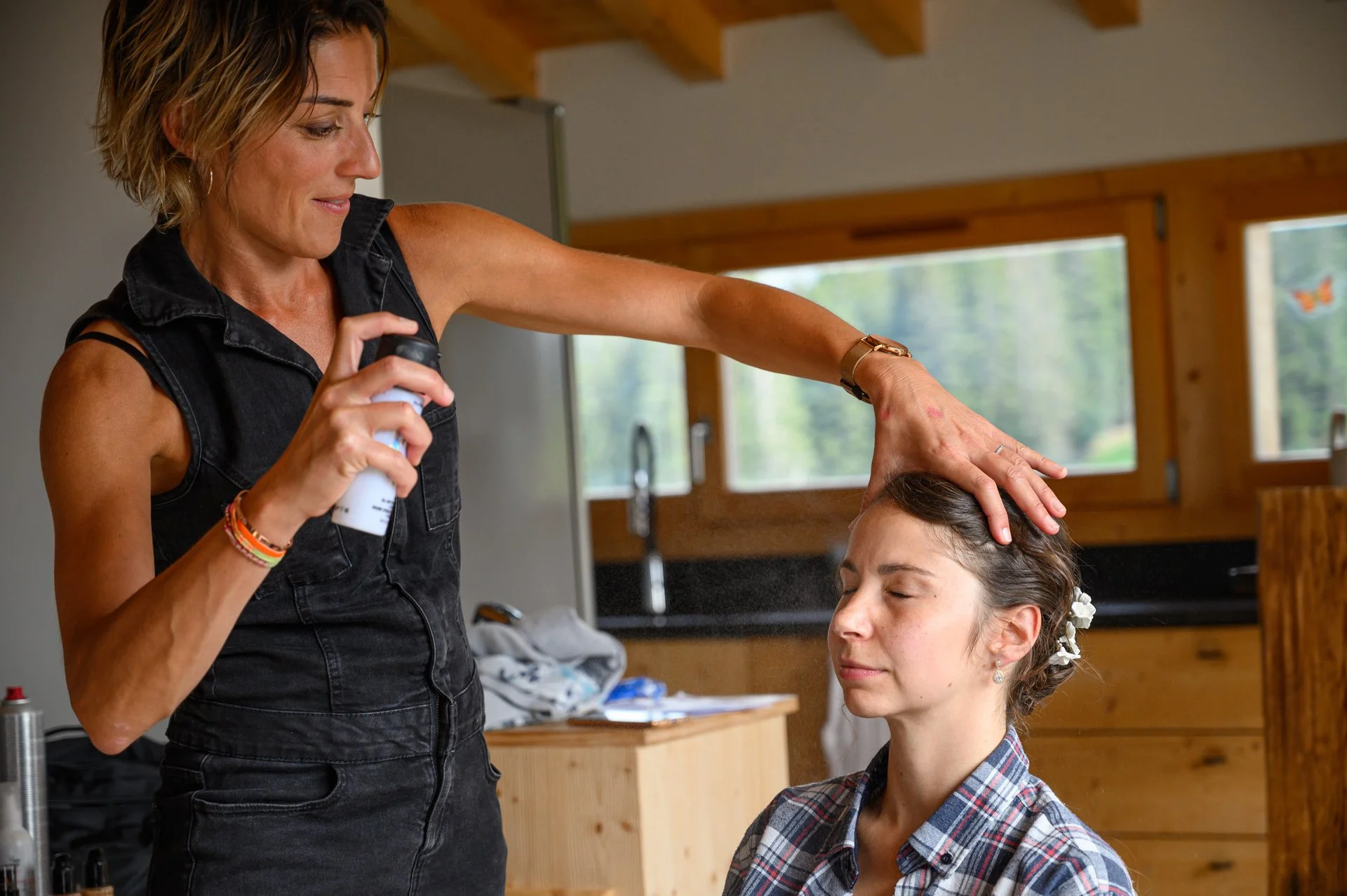Une femme prépare un spray sur une autre femme qui est assise, dans une pièce avec fenêtres en bois et vue sur la nature.
