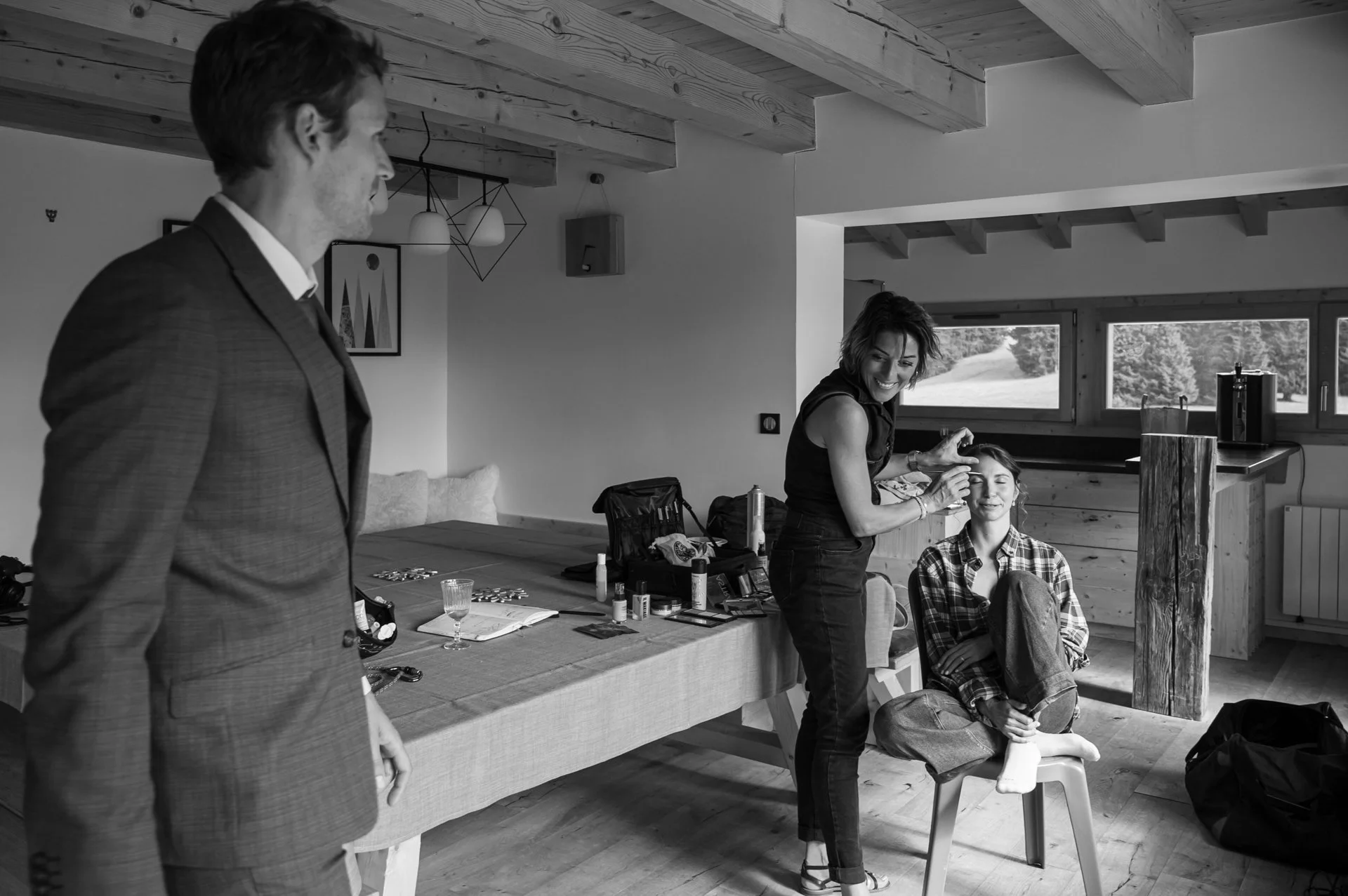 Une femme fait des soins du visage ou du maquillage à une femme assise sur une chaise dans une pièce avec de grandes fenêtres, un homme en costume à gauche observe.