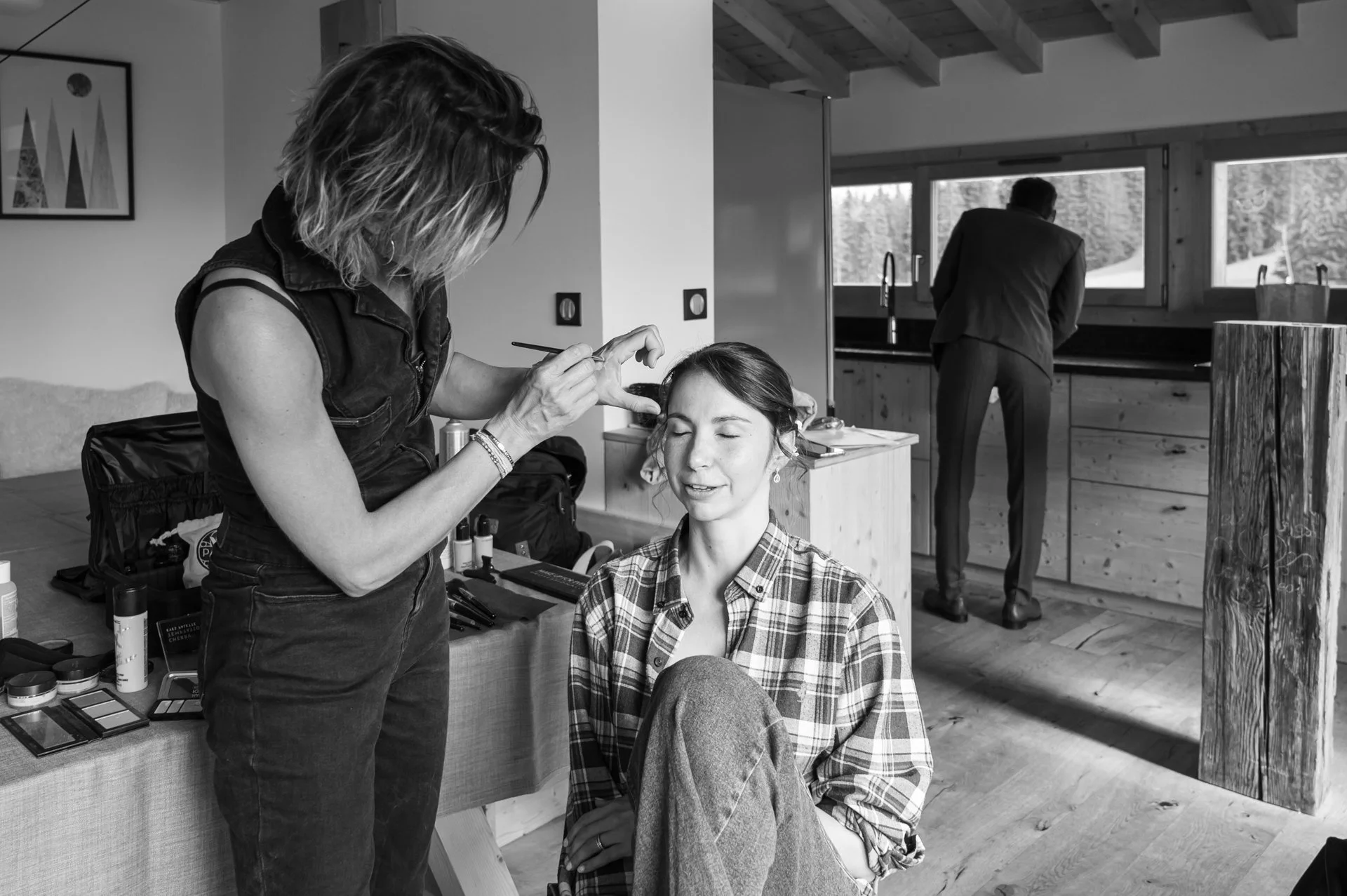 Une maquilleuse fait une séance de maquillage à une femme assise, alors qu'un homme se tient au comptoir de la cuisine en arrière-plan.