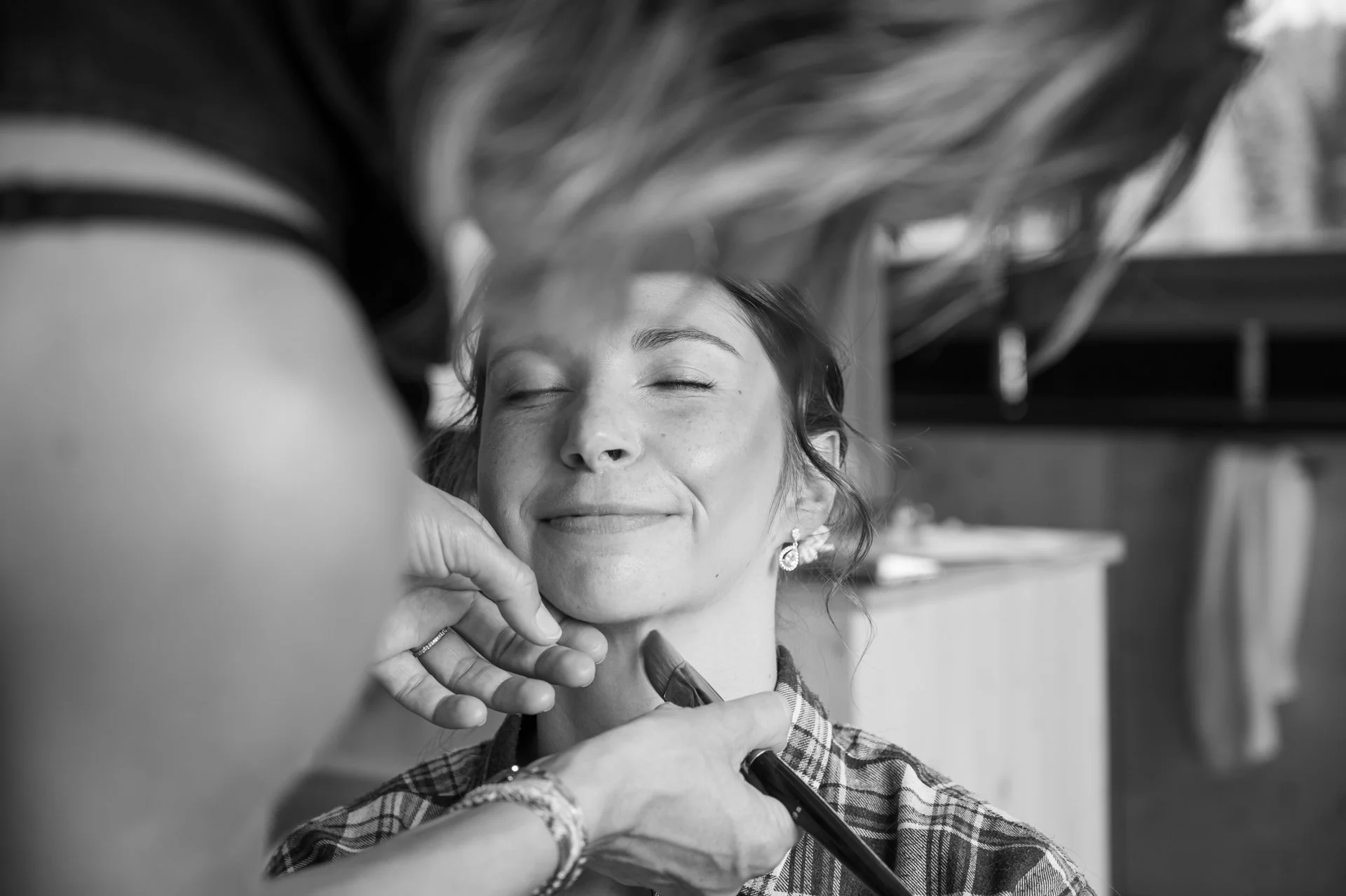 Une femme souriante a les yeux fermés pendant qu'une autre personne applique du maquillage sur son visage, dans un environnement intérieur.