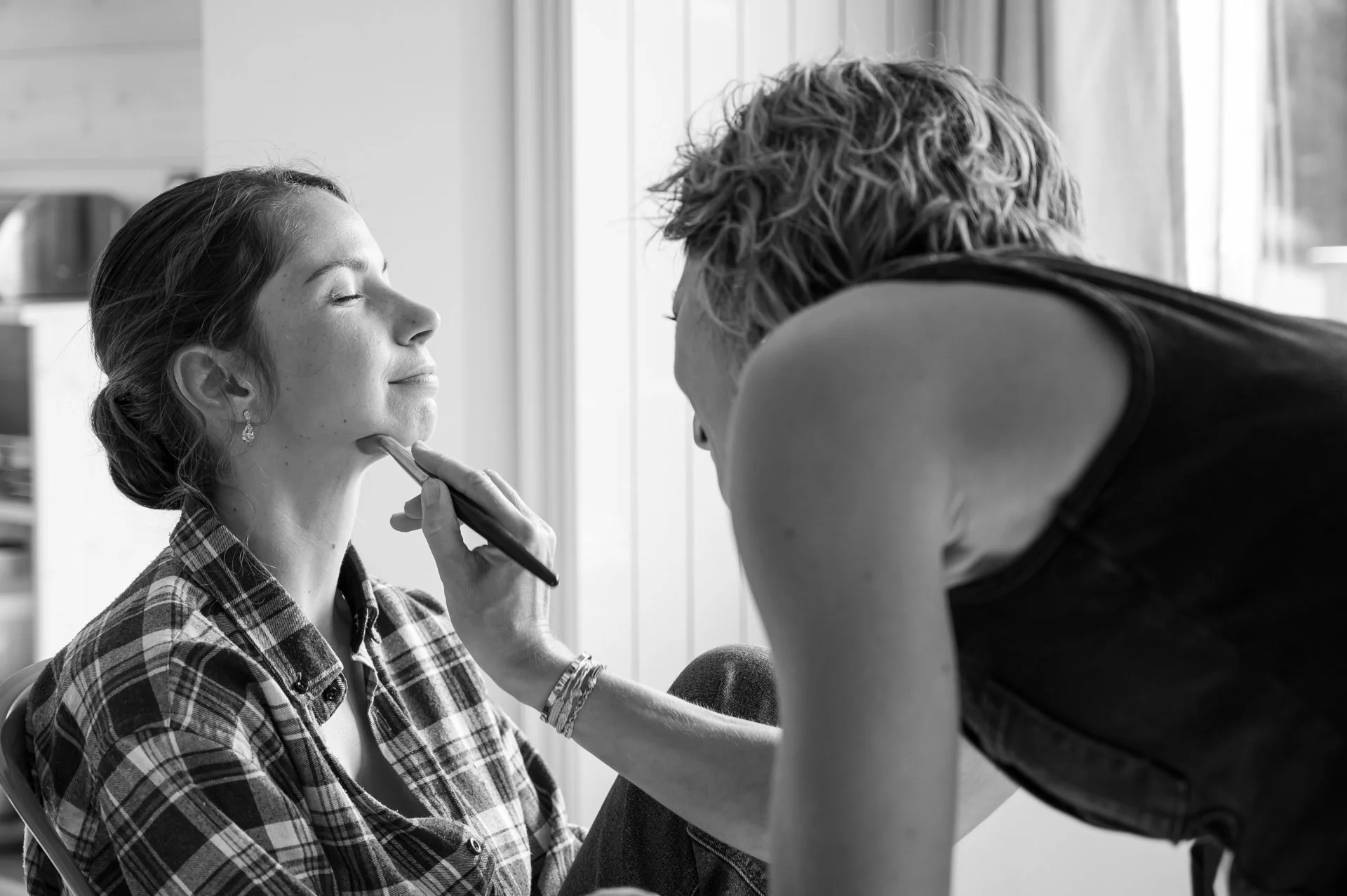 Une femme reçoit un maquillage, les yeux fermés, par une autre personne dans un cadre intérieur.