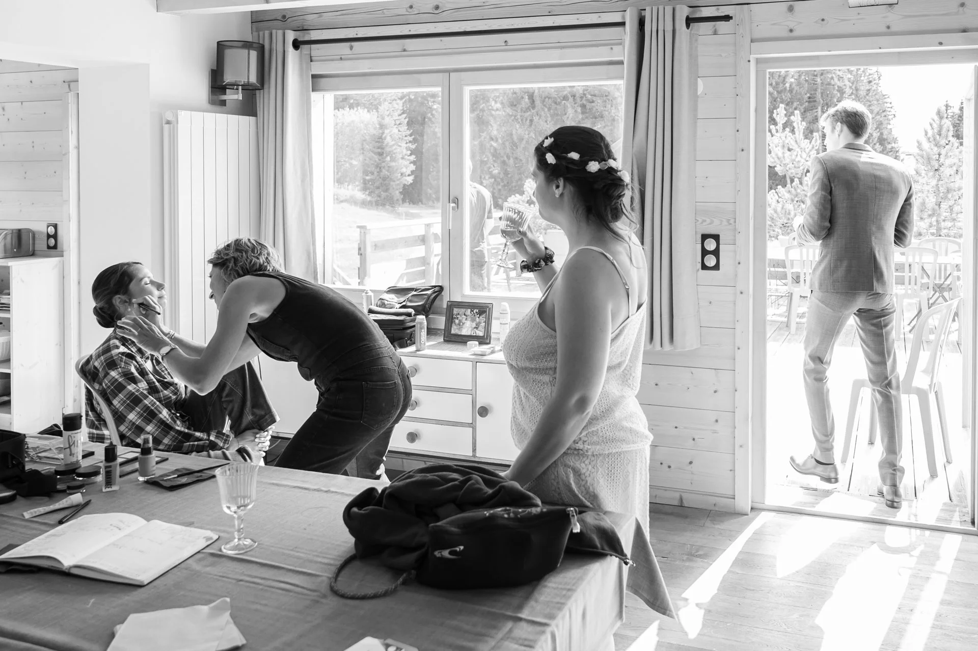 Scène intérieure d'une maison en bois où une femme se fait maquiller, une autre femme se prépare avec une boisson à la main, et un homme en costume se tient à l'extérieur sur un balcon, ensoleillé.
