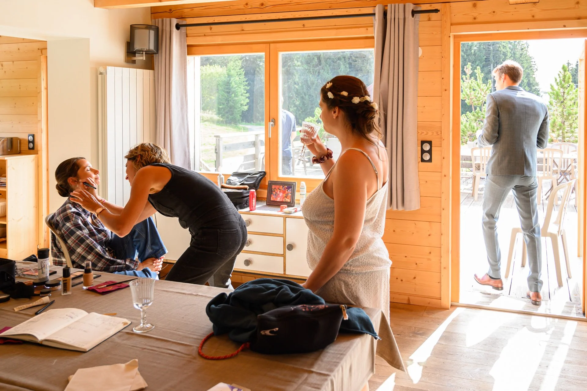 Femme en robe de mariée avec un verre, maquilleuse en train de faire le maquillage d'une autre femme, homme en costume qui sort de la maison par la porte à l'extérieur, dans une pièce en bois avec vue sur la nature.