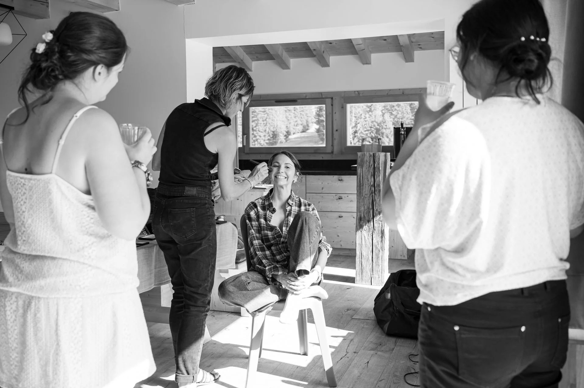 Une séance de maquillage ou de soins de beauté avec une femme souriante assise, entourée de trois autres femmes tenant des contenants ou des outils dans une pièce lumineuse en bois.