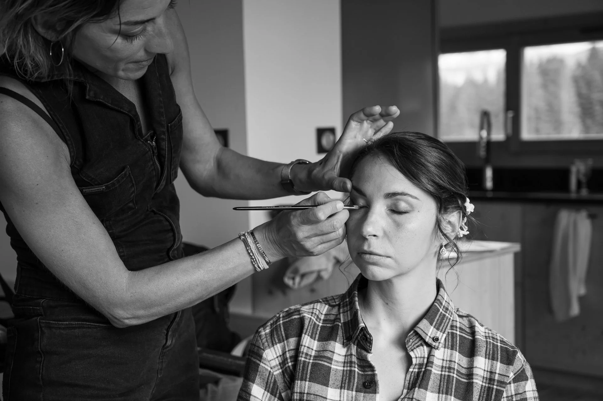 Une femme en train de faire du maquillage à une autre femme, dans une pièce avec une fenêtre. La femme maquilleuse utilise un pinceau, la femme se fait faire un maquillage des yeux.