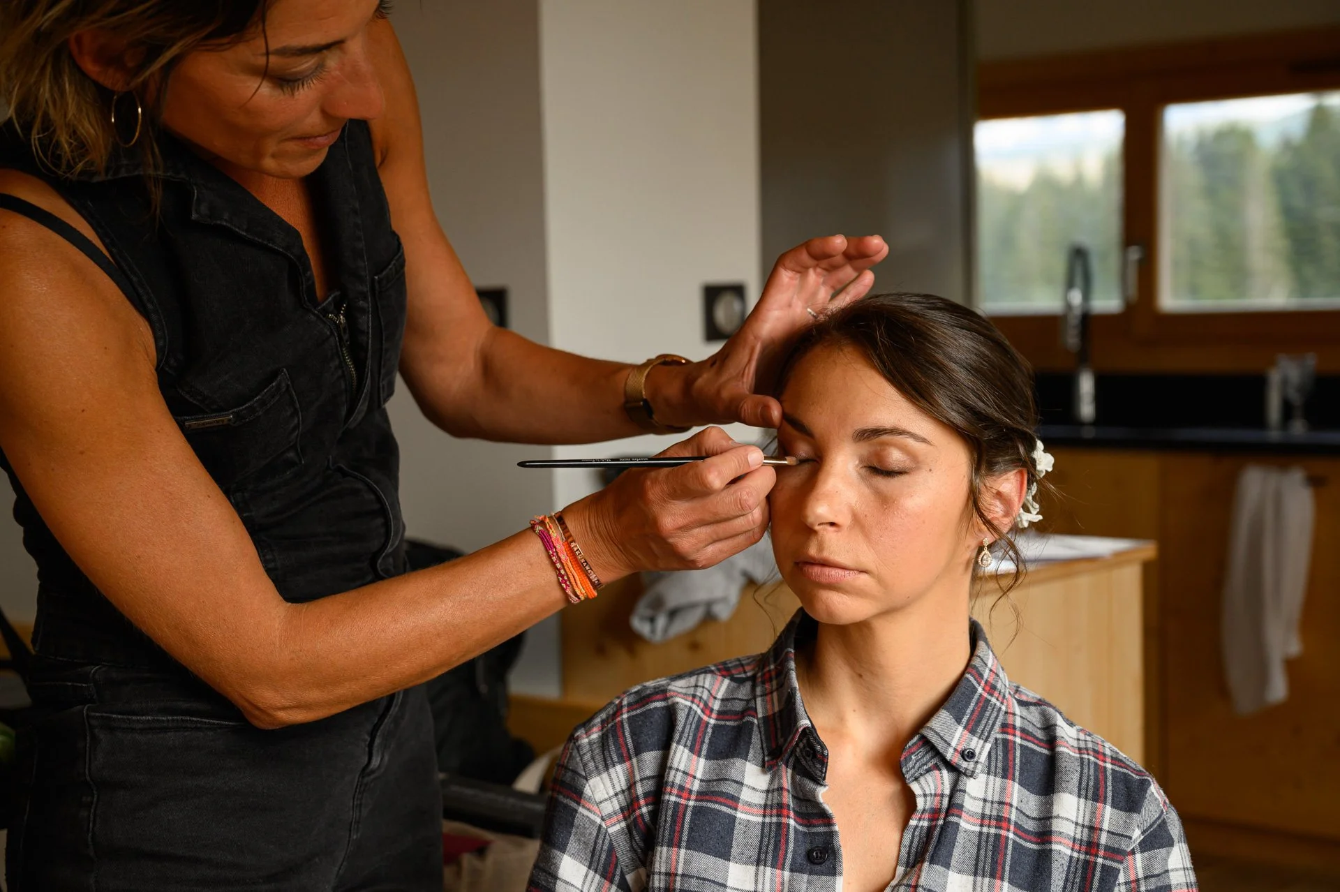 Une maquilleuse applique du maquillage sur les yeux d'une femme assise, dans une pièce bien éclairée.