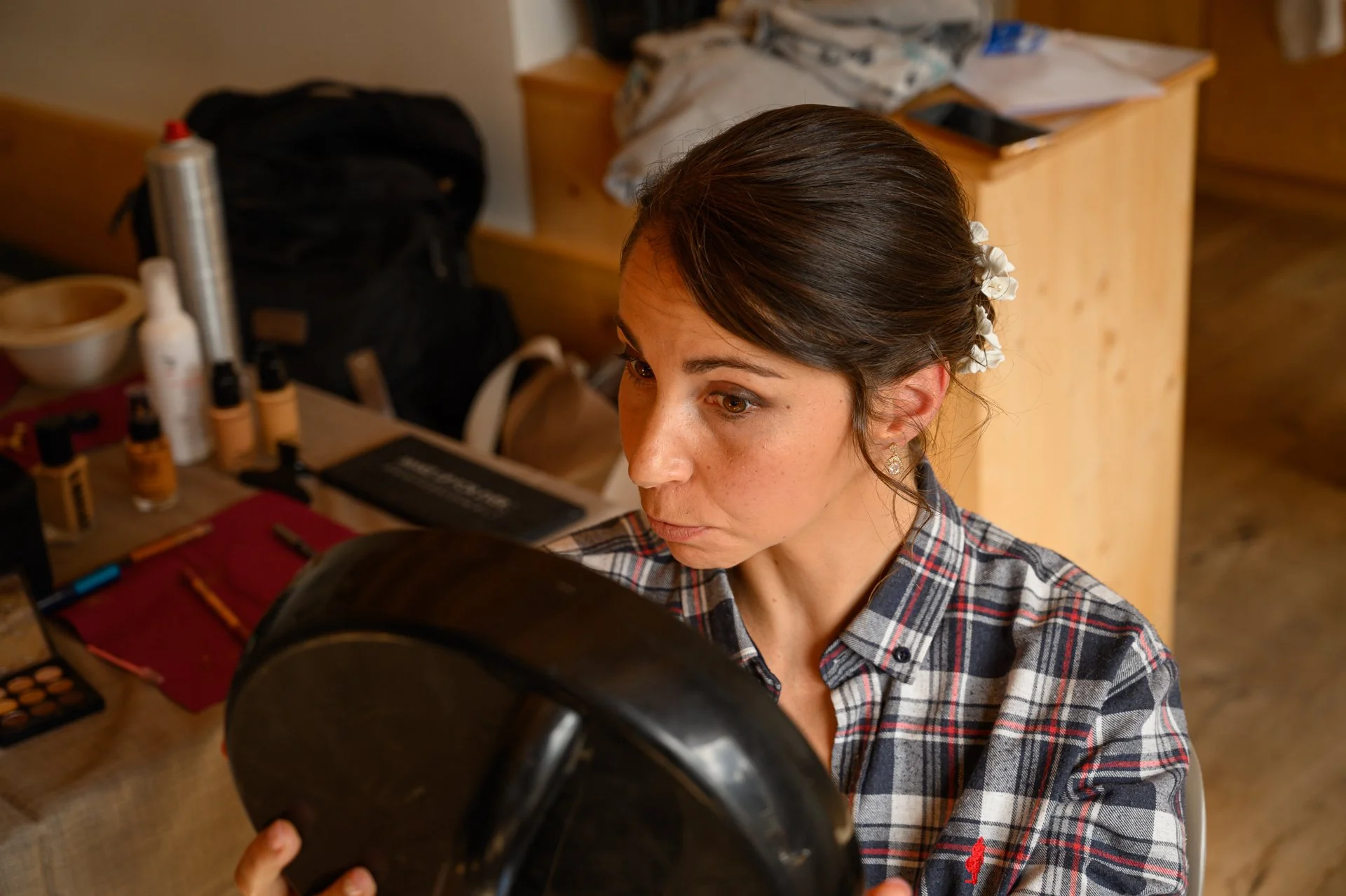 Femme regardant dans un miroir, portant une chemise à carreaux, avec des produits de maquillage et des objets sur la table à côté.