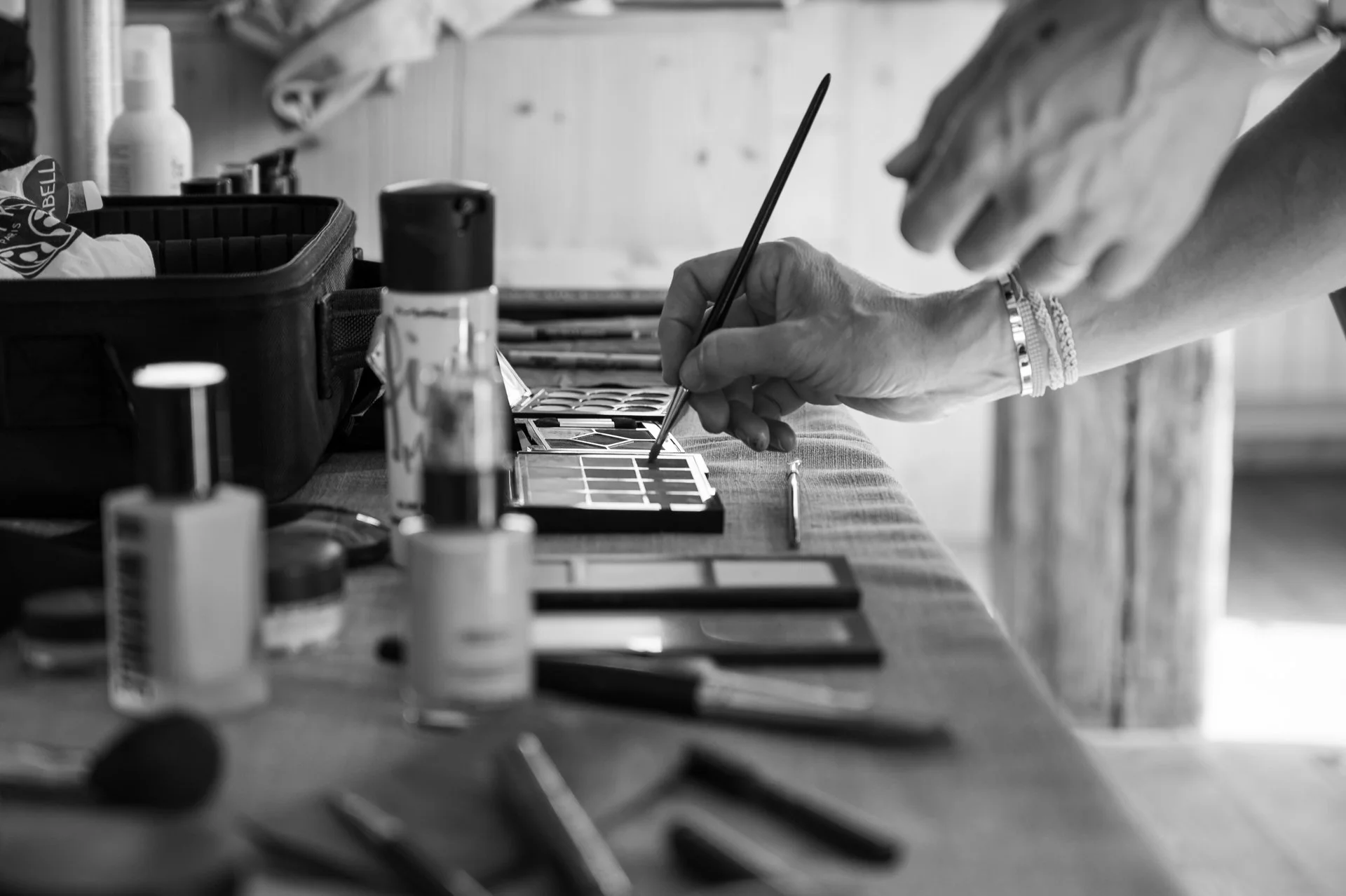 Une personne utilise un pinceau pour peindre ou appliquer du maquillage sur une palette contenant différentes nuances de couleurs, avec divers tubes et boîtes de peinture ou d'accessoires à proximité.