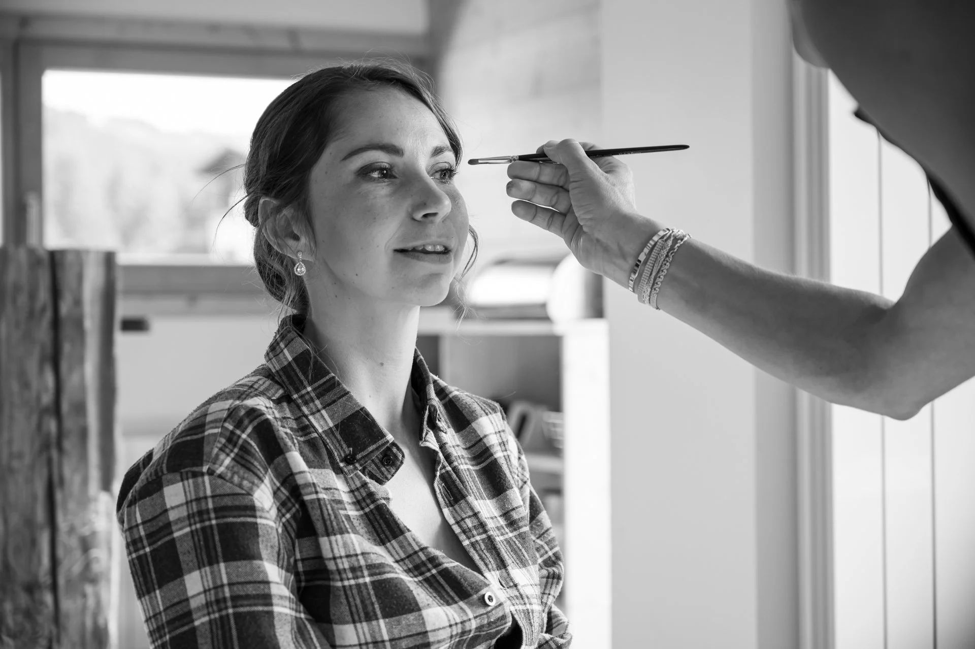 Une femme est en train de se faire maquiller, assise devant une fenêtre, portant une chemise à carreaux.