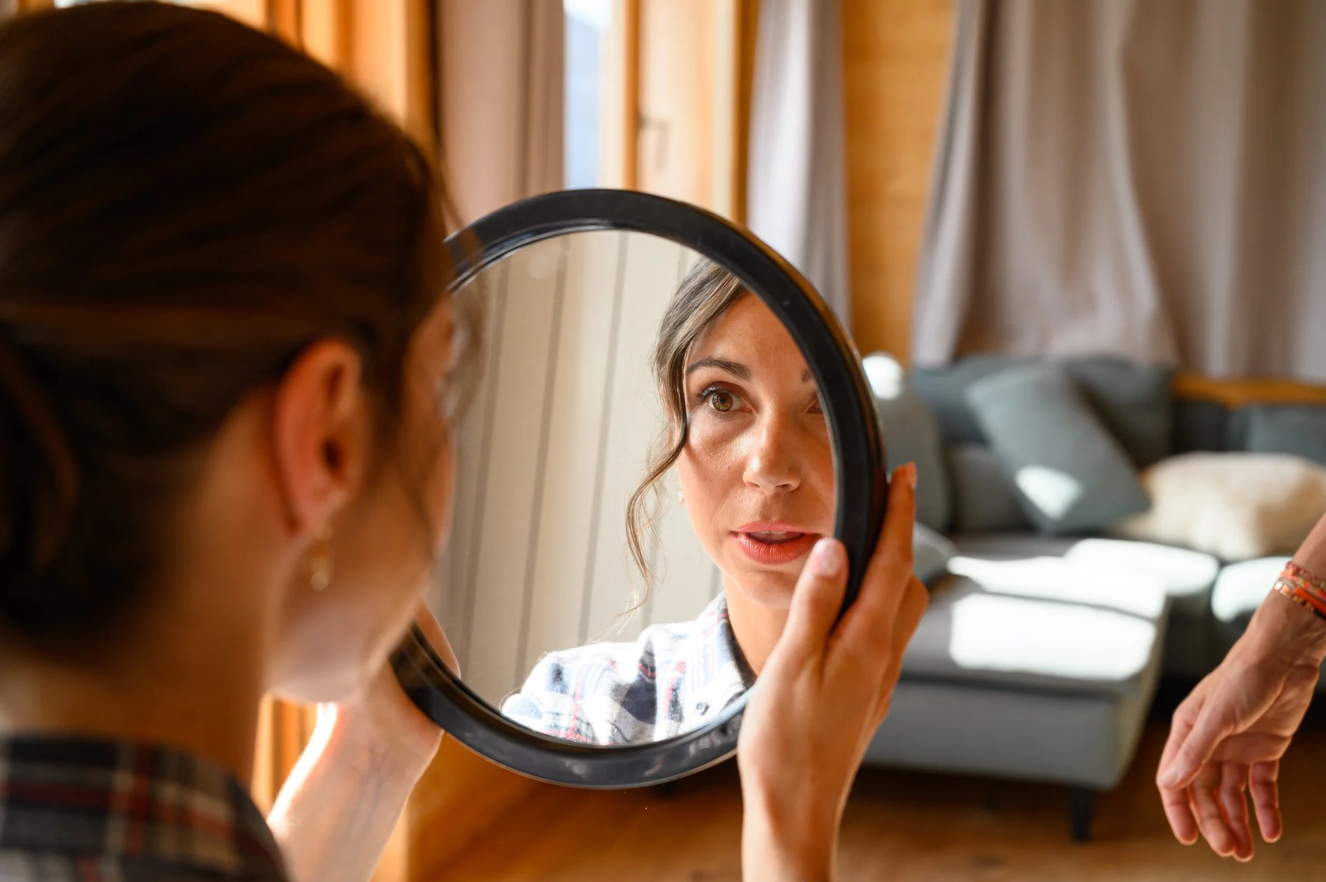 Une femme se regarde dans un miroir rond, tenant le miroir devant son visage, dans une pièce lumineuse avec un canapé en arrière-plan.