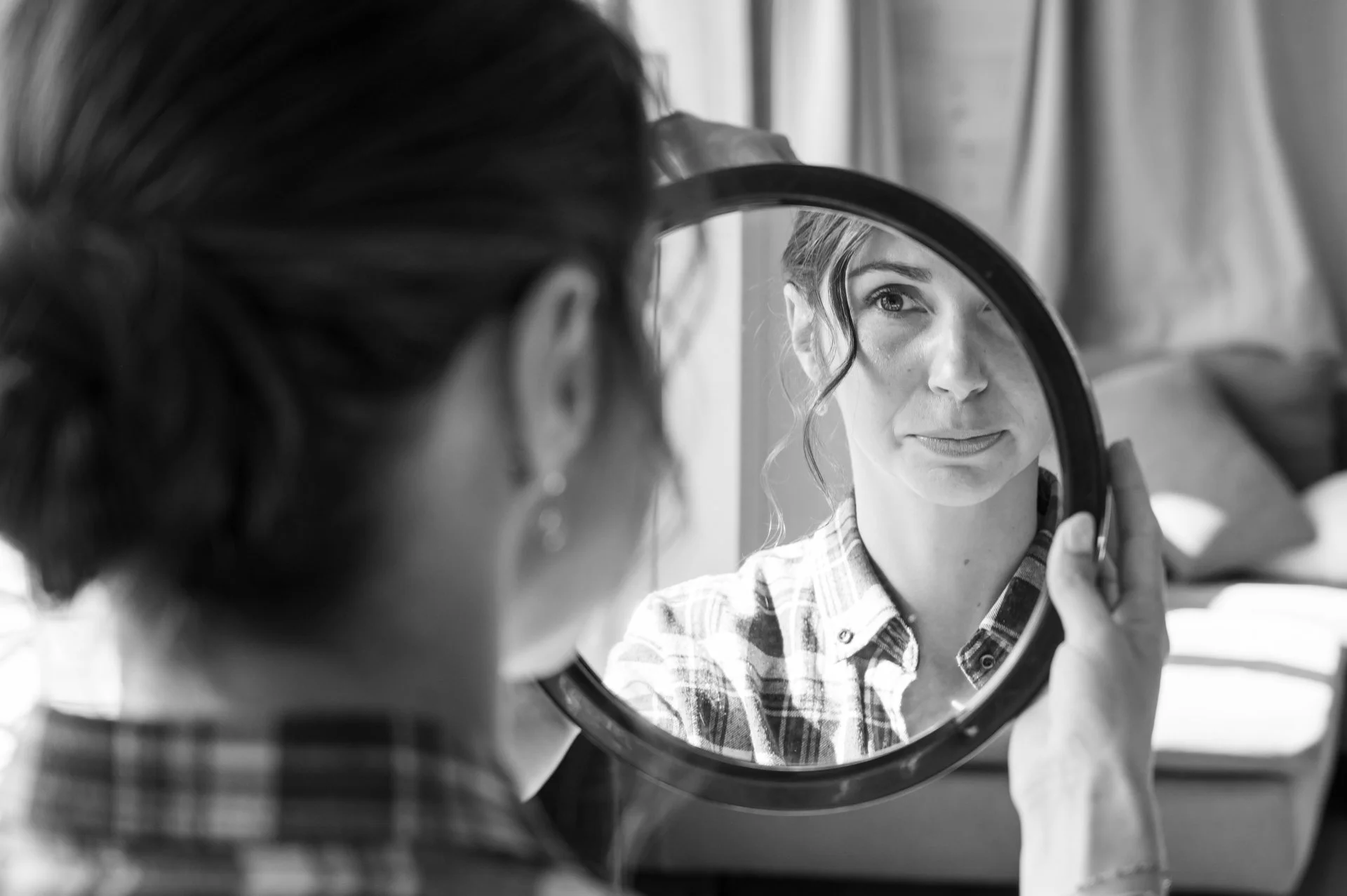 Une femme regarde dans un miroir rond, reflétant son visage avec expression sérieuse, portant une chemise à carreaux, dans un intérieur domestique.