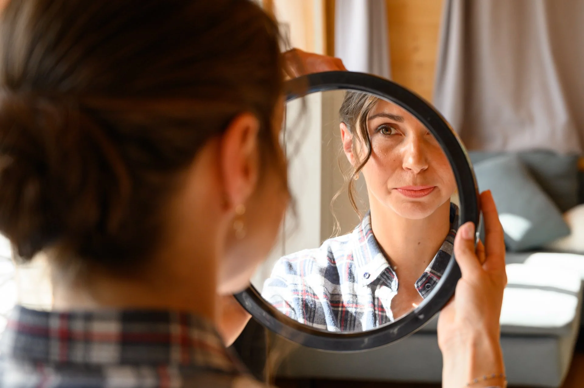 Une femme se regarde dans un miroir circulaire, vêtue d'une chemise à carreaux, dans une pièce lumineuse.