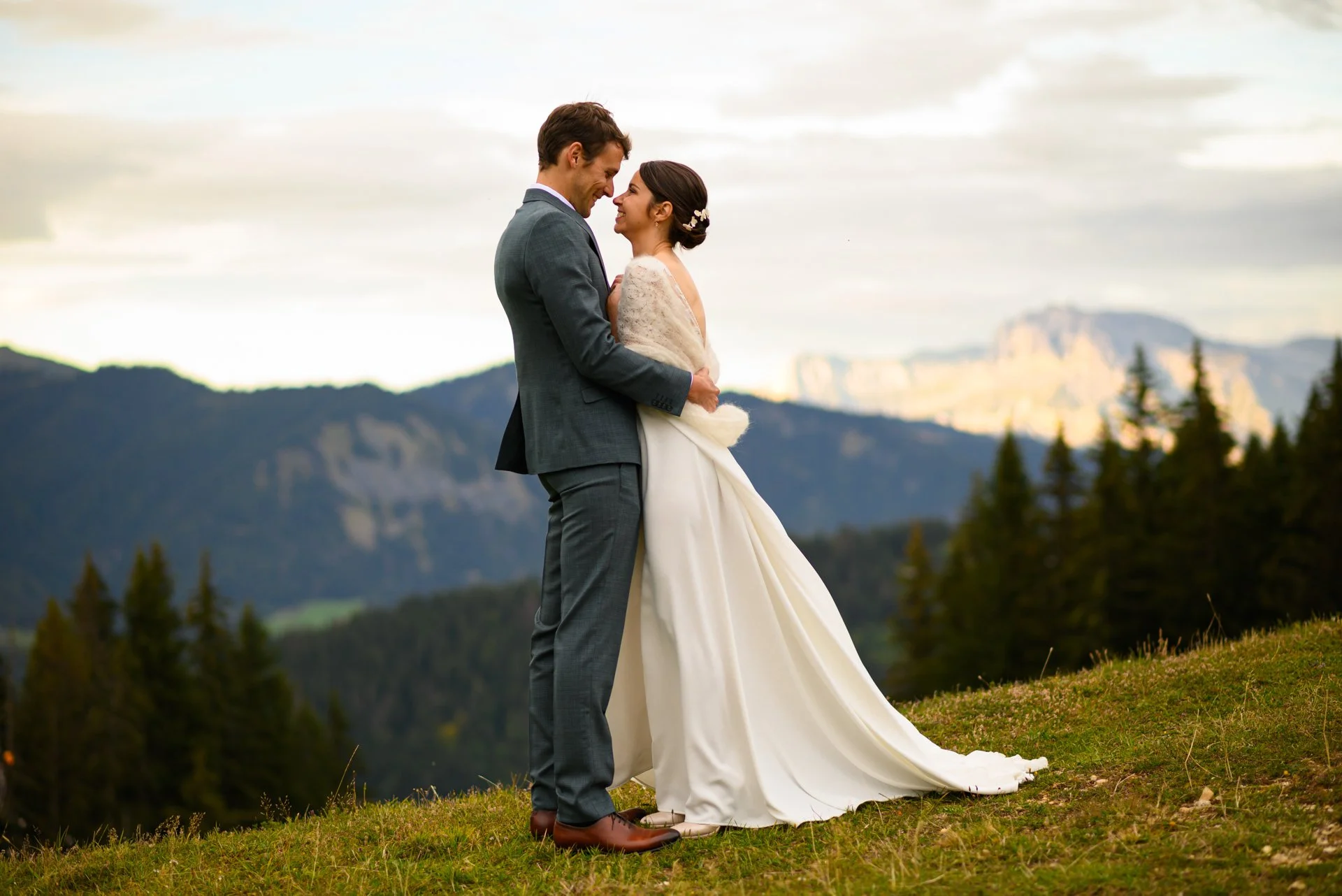 Un couple de mariés se tient dans un paysage de montagne, en regardant joyeusement. La femme porte une robe de mariée blanche et le homme un costume gris. Il y a des collines et des montagnes en arrière-plan, sous un ciel nuageux.