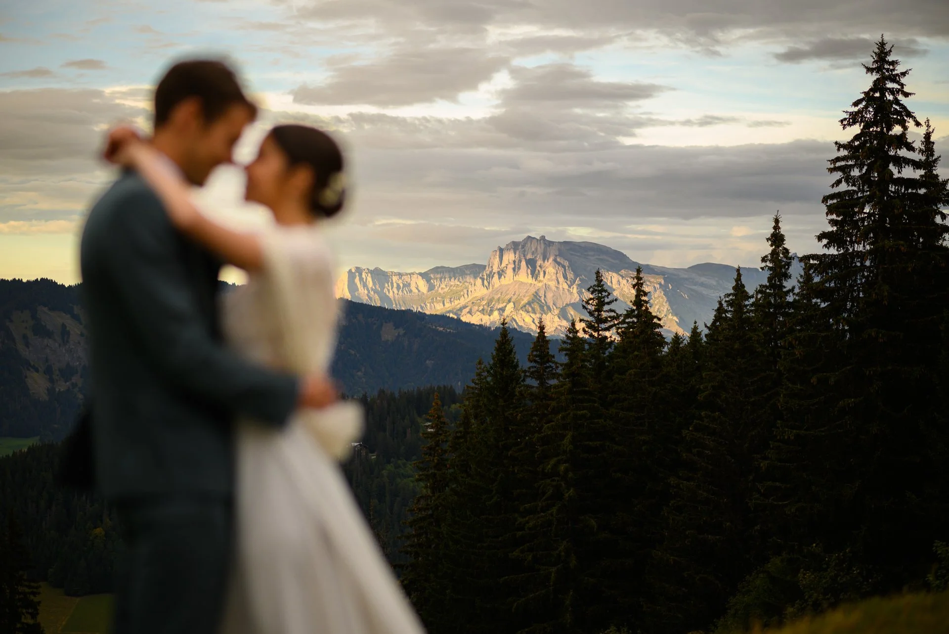Un couple de mariés nouvellement mariés qui s'embrassent dans un paysage de montagne, avec des forêts et des montagnes en arrière-plan, lors d'un coucher de soleil.