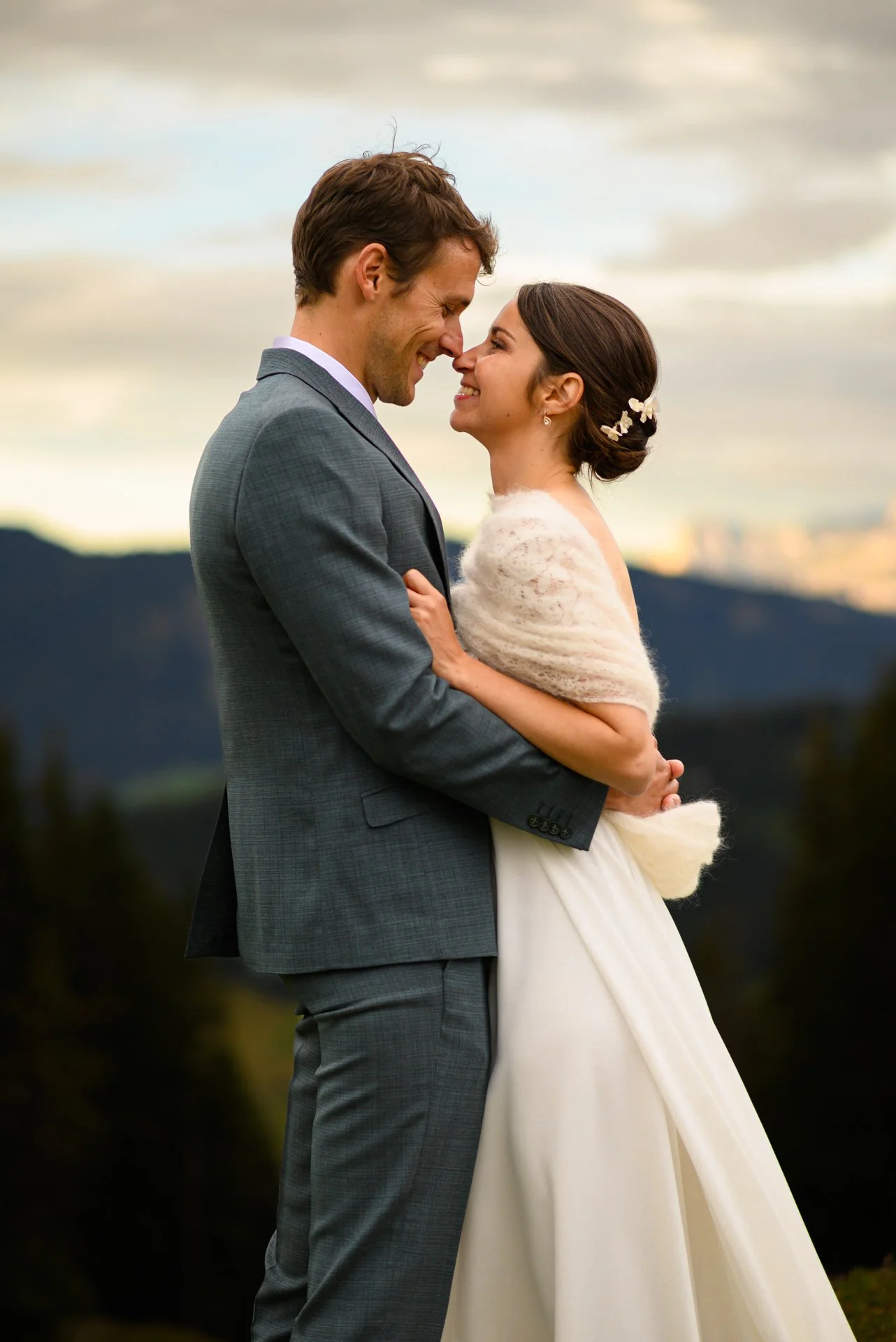 Jeune couple de mariés souriant, en tenue de mariage, se tenant très proches, dans un paysage de montagnes au coucher du soleil.