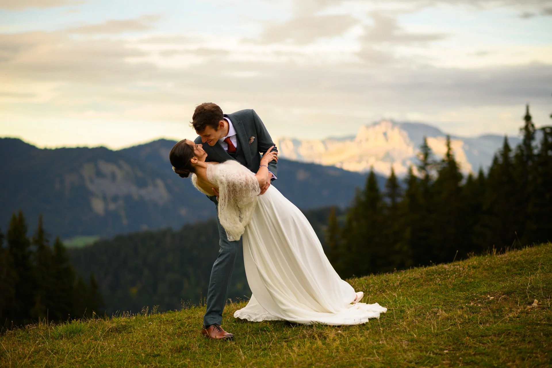 Un couple de mariés danse dans un champ avec des montagnes en arrière-plan, lors du coucher du soleil.