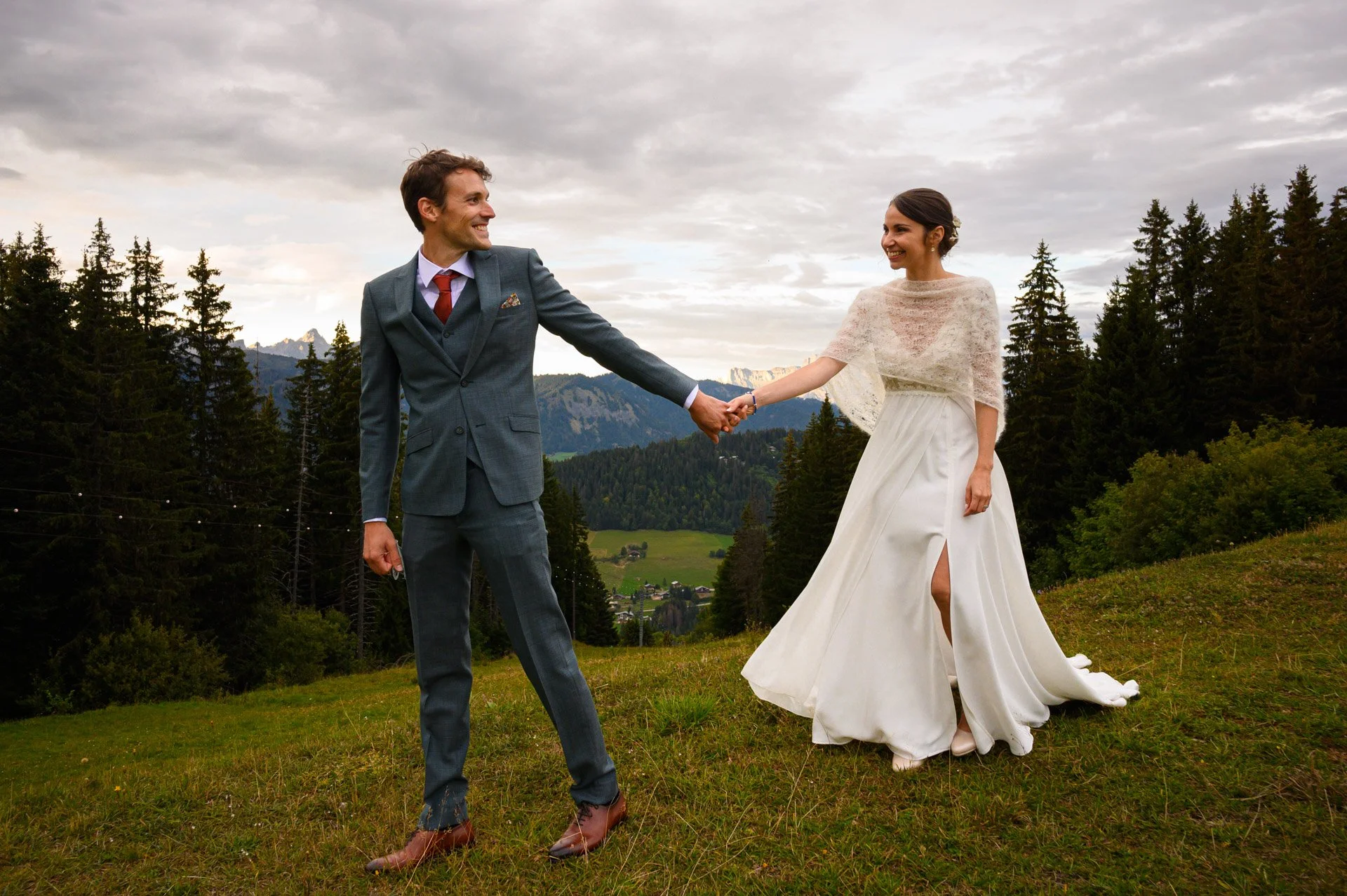 Un couple de mariage dans un champ vert, avec des montagnes et un ciel nuageux en arrière-plan, la femme porte une robe blanche et l'homme un costume gris.