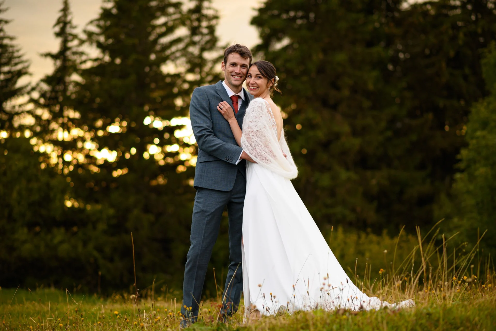 Un couple de mariés souriant et posant dans un champ avec des arbres en arrière-plan lors d'un coucher de soleil.