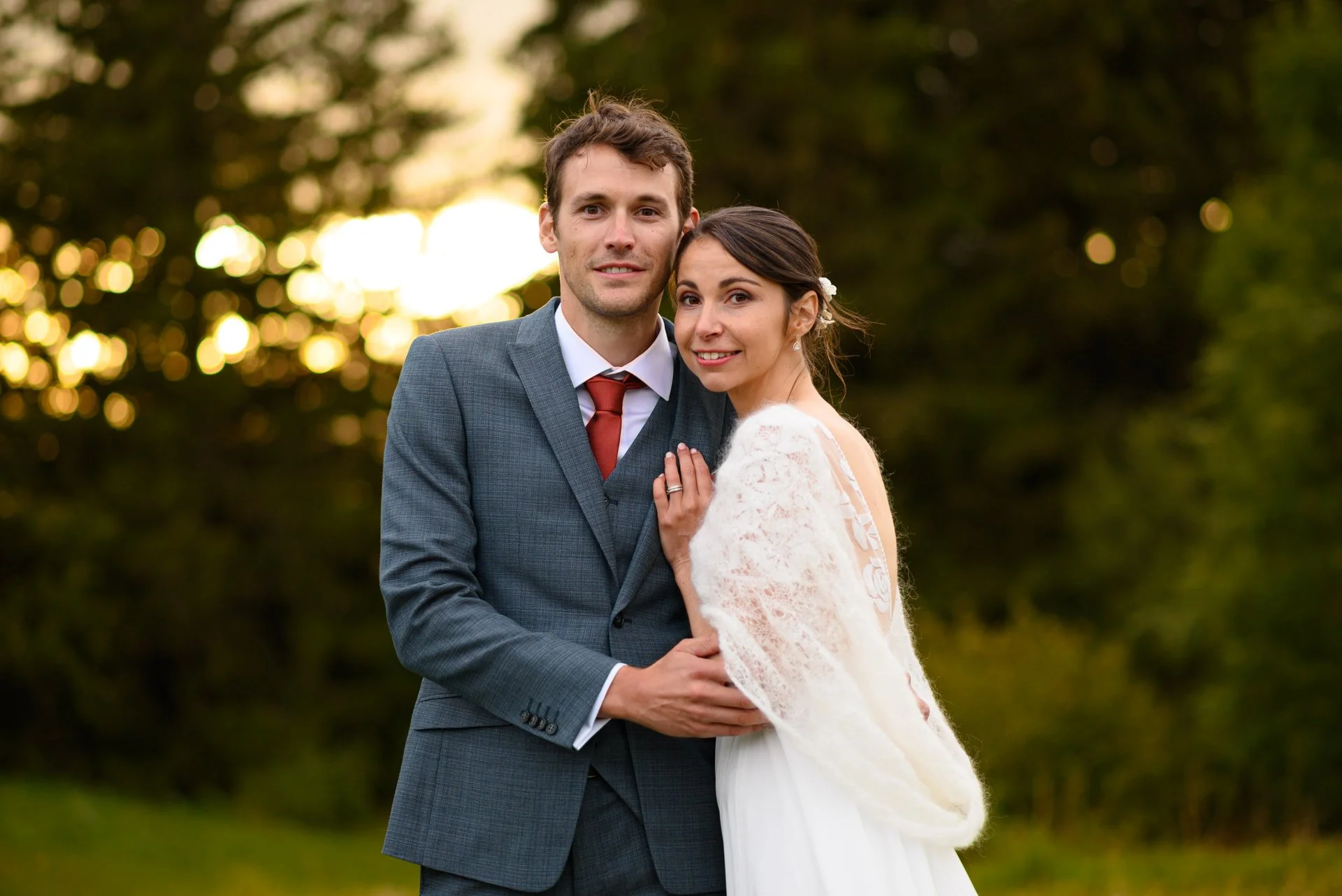 Un couple en tenue de mariage, le homme en costume gris et la femme en robe blanche, se tenant dans un parc lors du coucher du soleil.