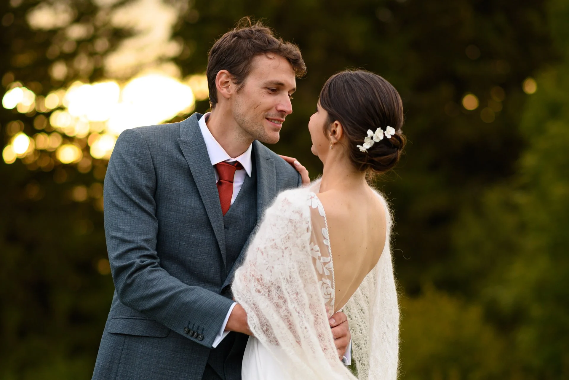 Un couple de mariés en plein air lors d'une séance photo de mariage, l'homme en costume gris et la femme en robe blanche avec des fleurs dans les cheveux, se regardant tendrement.