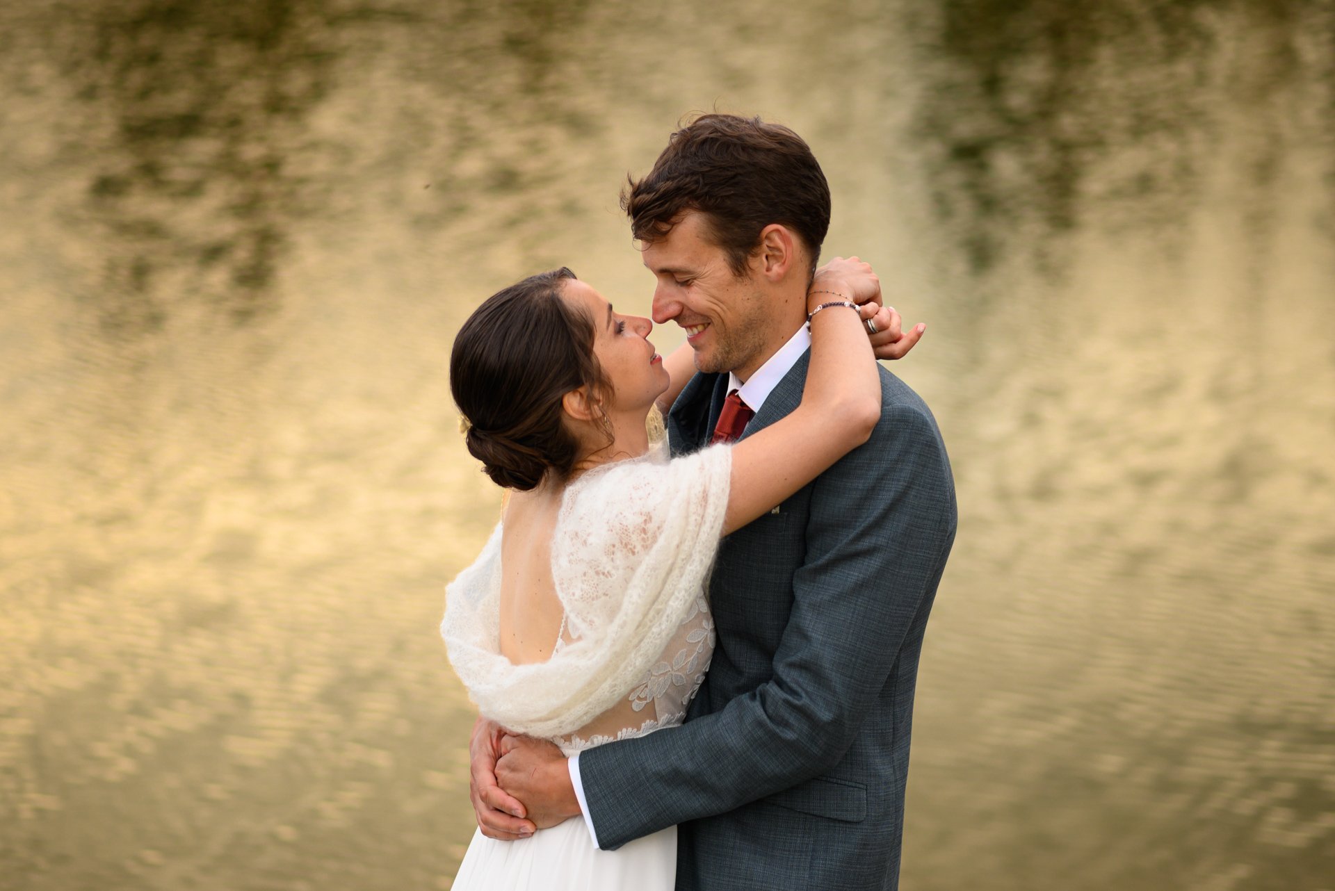 Un couple embrassé devant un lac, l'homme porte un costume gris et la femme une robe blanche avec un châle.