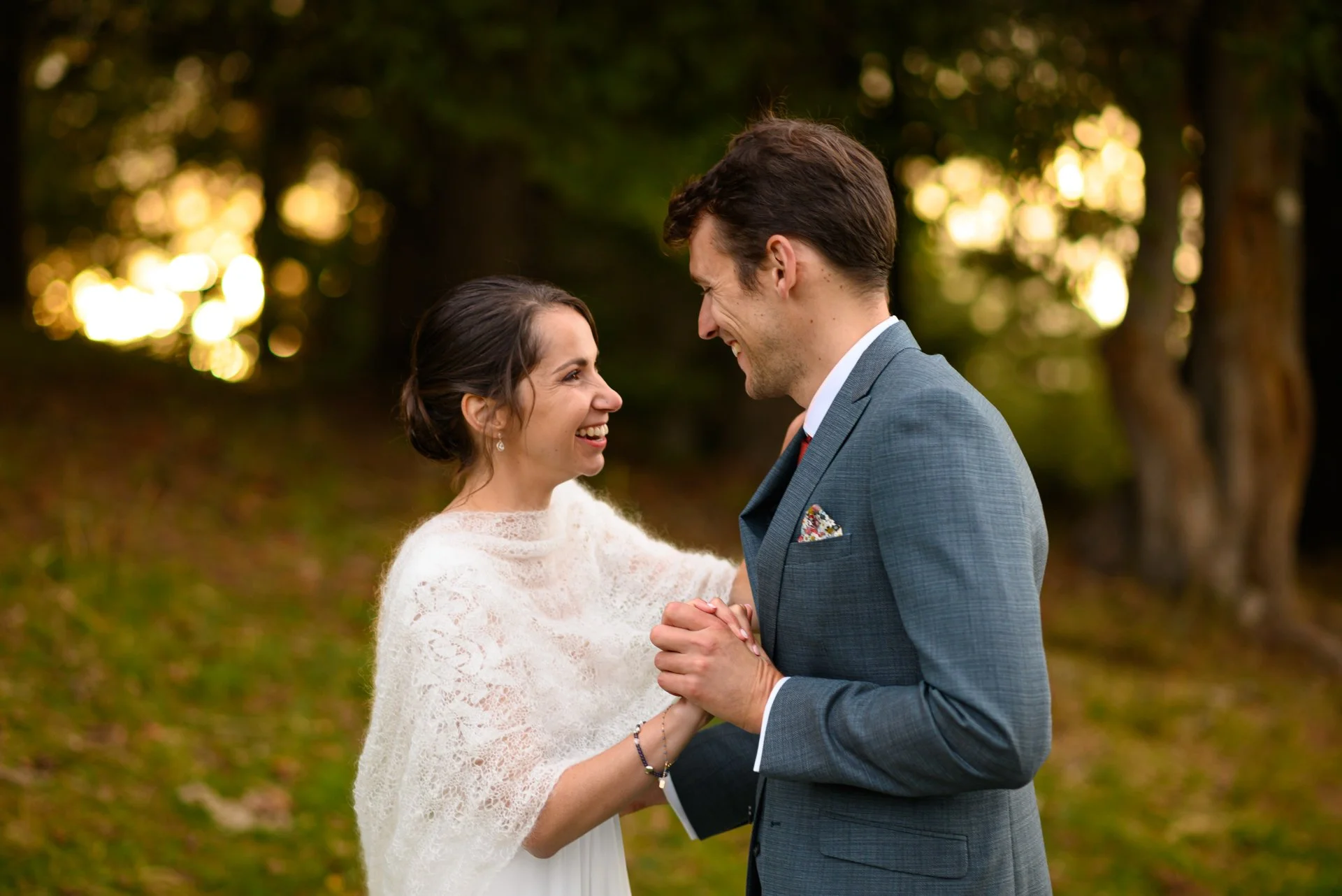 Un couple souriant, la femme en robe blanche avec une étole en lin, l'homme en costume gris, se tenant la main dans un parc avec des arbres en arrière-plan pendant un coucher de soleil.